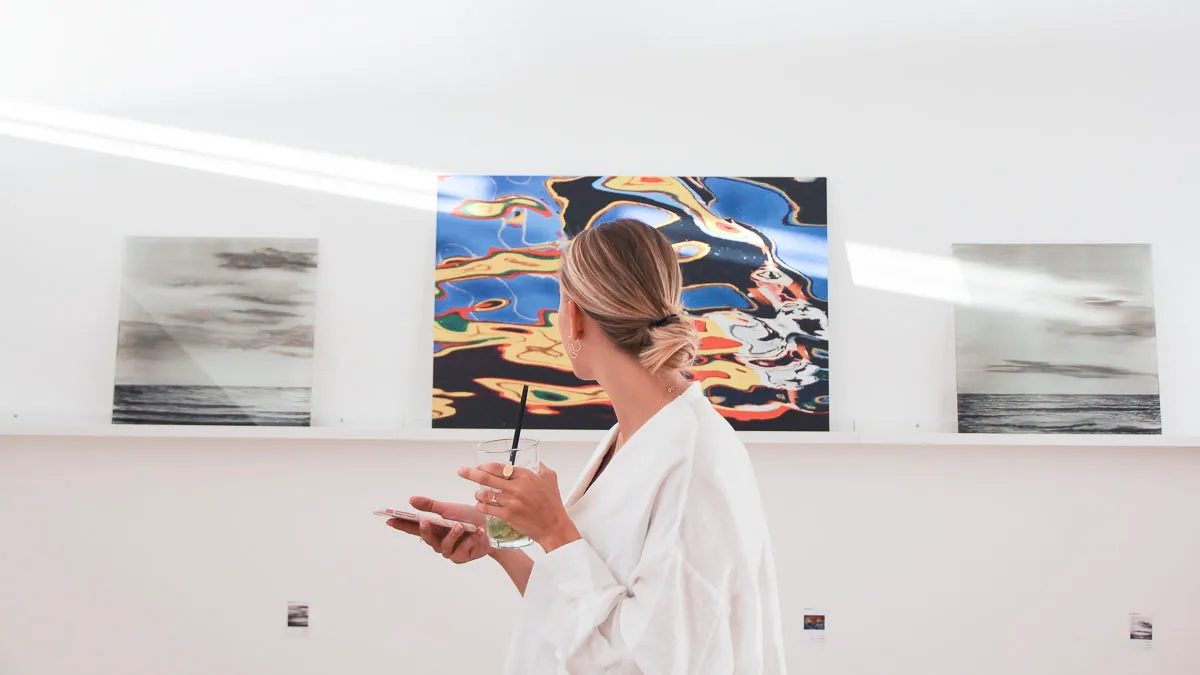Una mujer con una túnica blanca que sostiene una paleta y un pincel está de pie frente a tres pinturas en la pared blanca de una galería, con una obra abstracta y colorida centrada entre dos piezas en tonos grises.