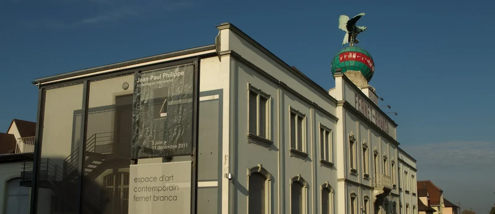 Biało-szara fasada budynku ze skrzydlatym posągiem na globusie na dachu oraz dużym banerem z boku z napisem musée d'art contemporain fernet branca