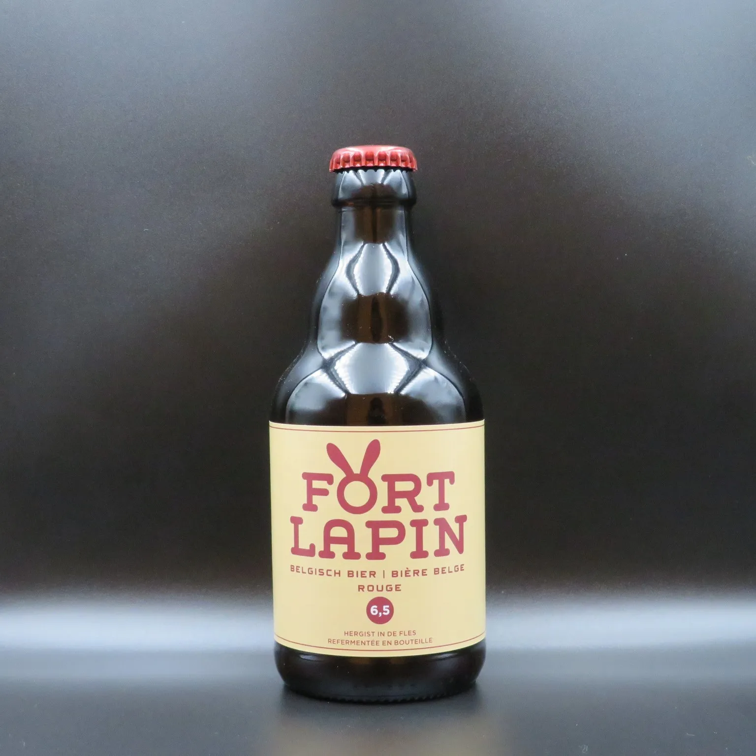 Una botella de cristal marrón de cerveza belga Fort Lapin Rouge con una etiqueta color crema que presenta un logotipo de conejo rojo y una tapa roja, centrada sobre un fondo gris oscuro.