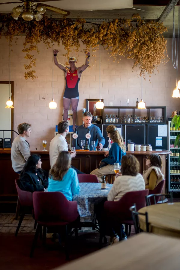 Interior de la sala de degustación de una cervecería donde un grupo de visitantes se sienta a una mesa y en la barra escuchando a un guía, bajo un gran mural de un forzudo sosteniendo un techo cubierto de lúpulo seco.