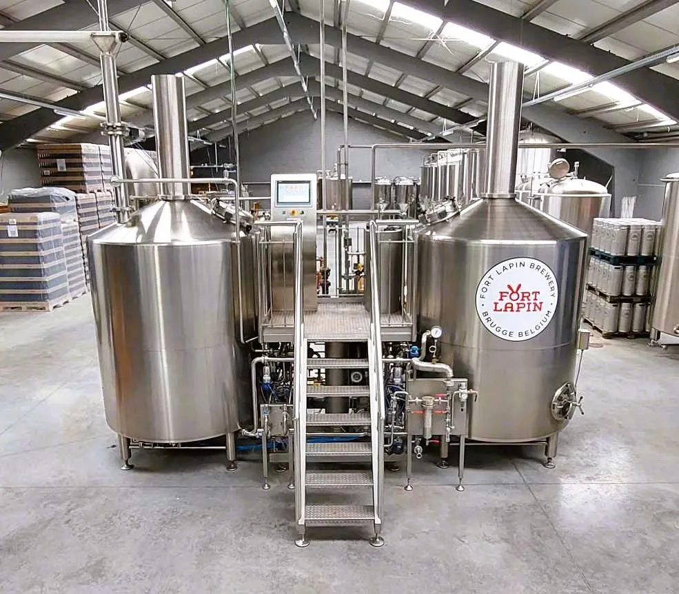 Interior de la cervecería Fort Lapin que muestra grandes tanques de fermentación de acero inoxidable con el logotipo de la cervecería visible en uno de ellos, pasarelas y escaleras metálicas entre los recipientes y barriles apilados al fondo.