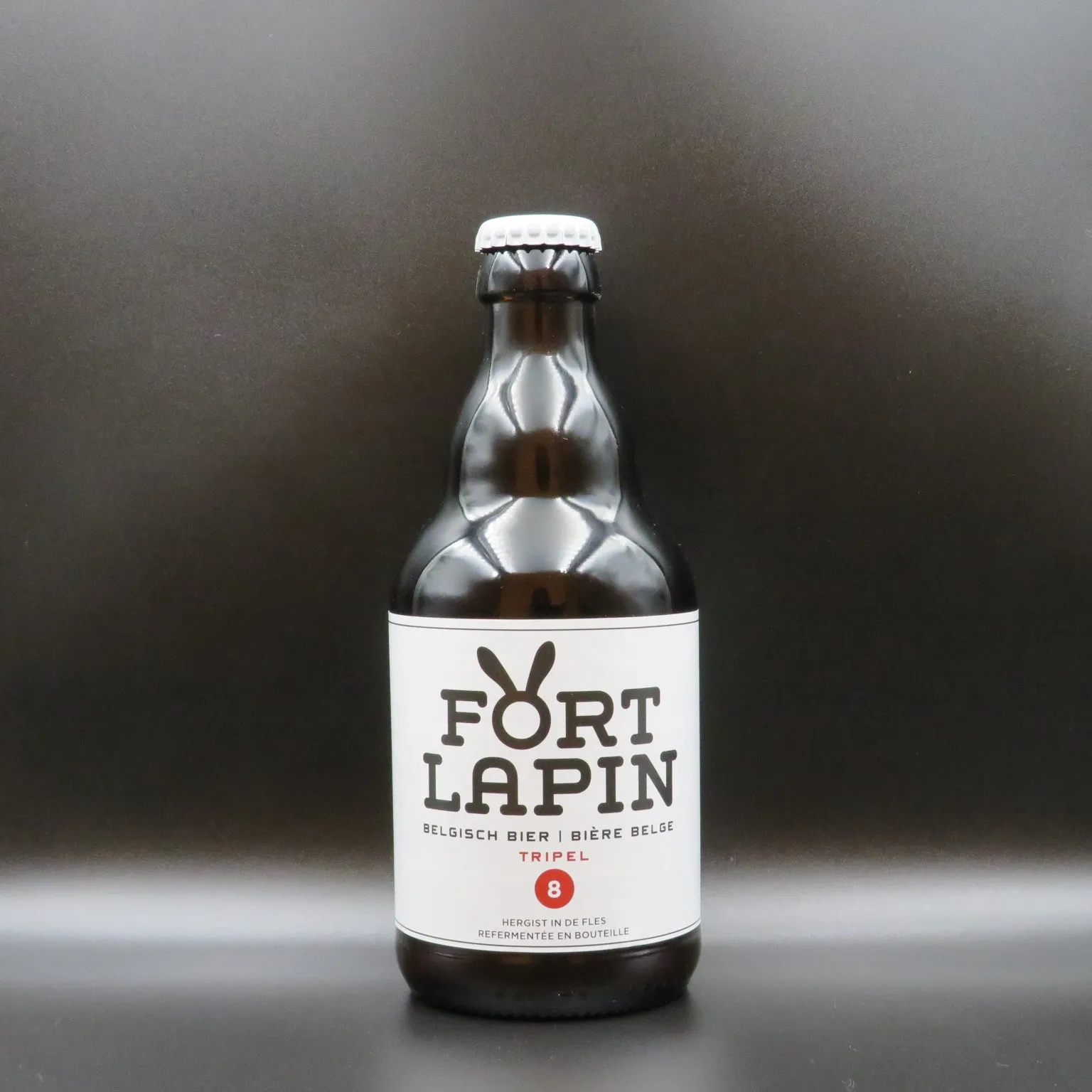 Una botella de cerveza de cristal marrón con una etiqueta blanca que dice FORT LAPIN y TRIPEL, con el logotipo de un conejo, sobre un fondo gris oscuro.