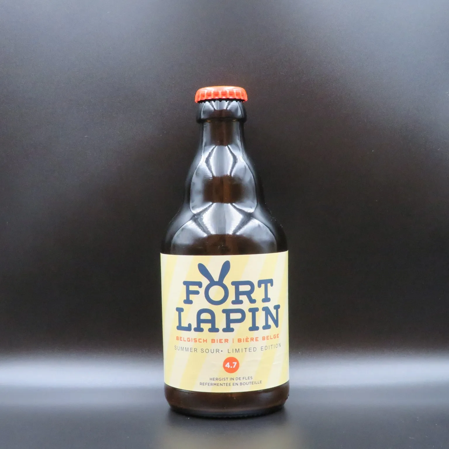 Una botella de vidrio marrón de cerveza belga Fort Lapin con etiqueta amarilla y tapón naranja, sobre un fondo gris oscuro.