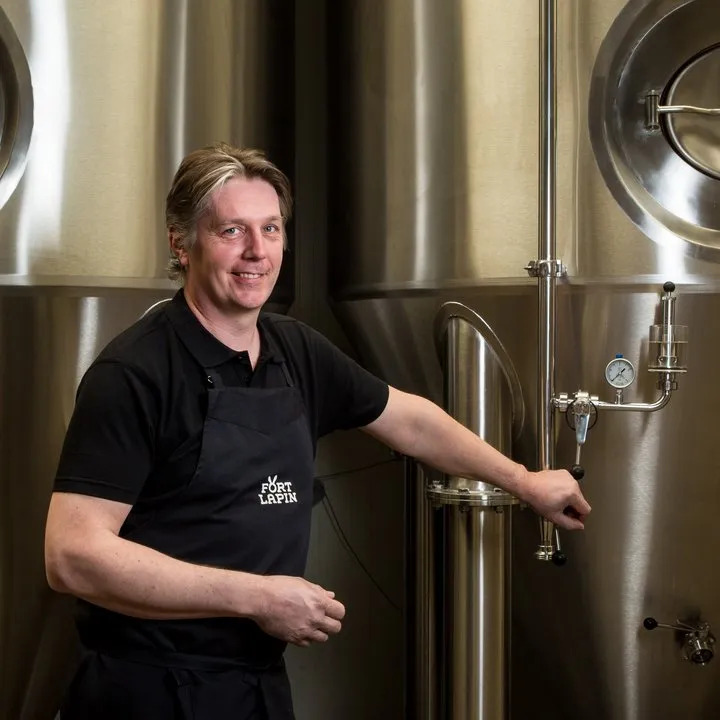 Un hombre sonriente que viste un delantal negro de Fort Lapin está de pie frente a grandes tanques de fermentación de acero inoxidable, apoyando su mano sobre una válvula.