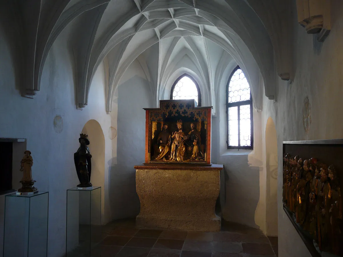 Gotycka sala muzealna ze sklepieniem, zawierająca średniowieczny rzeźbiony ołtarz drewniany na kamiennym postumencie, w otoczeniu rzeźb i łukowych okien.