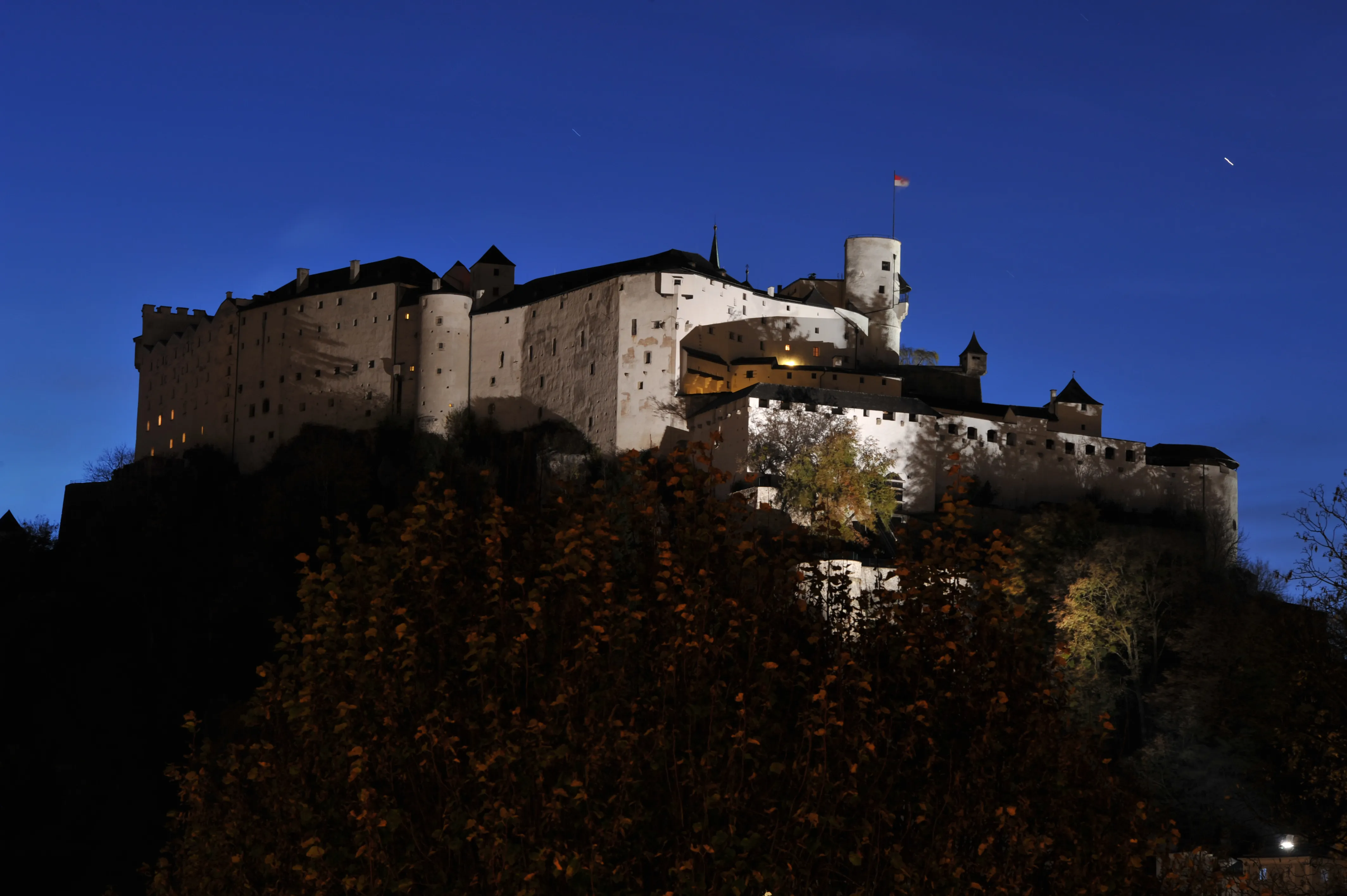 Twierdza Hohensalzburg oświetlona nocą na tle ciemnoniebieskiego nieba z jesiennymi drzewami na pierwszym planie