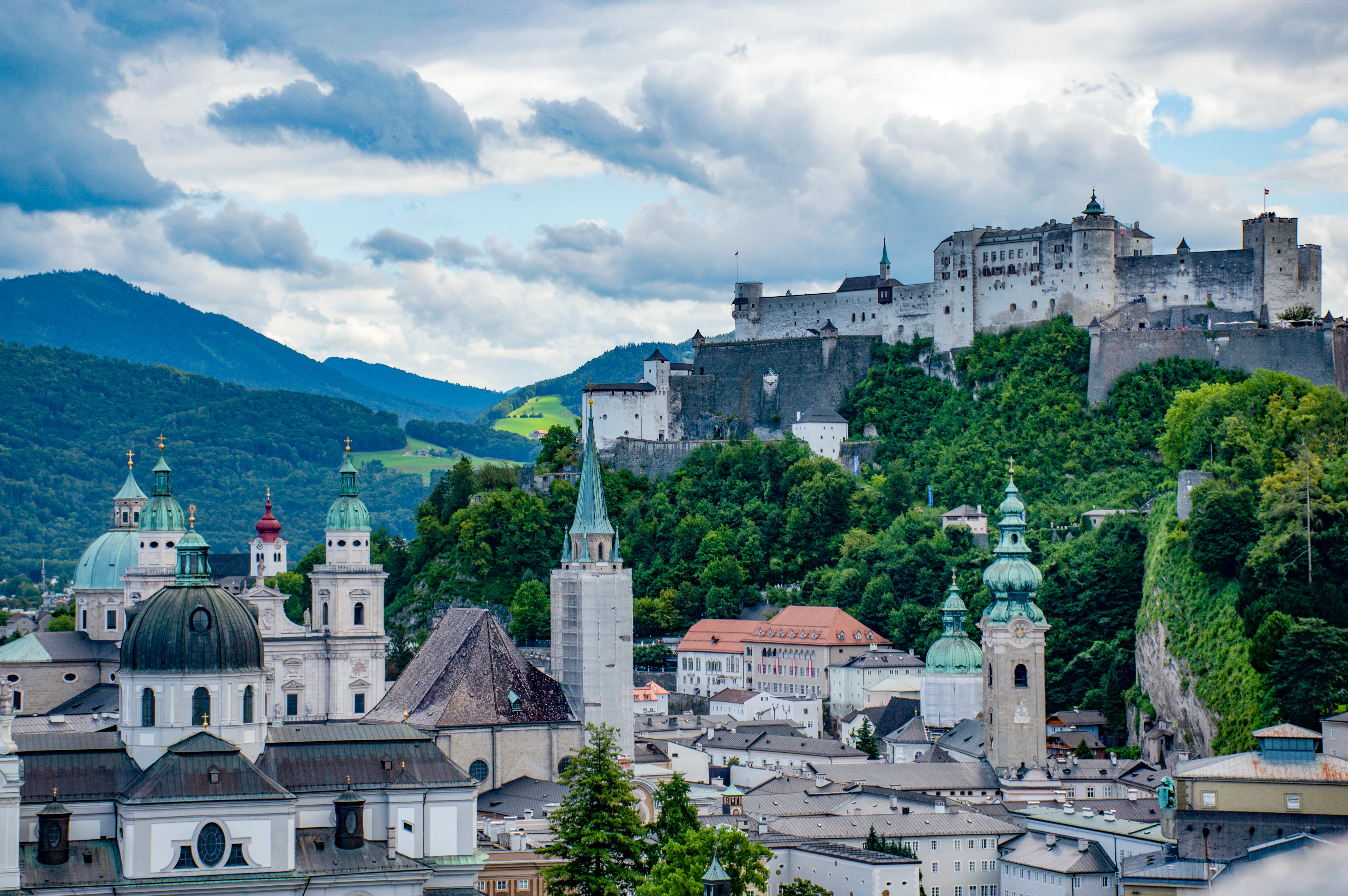 Panoramiczny widok na Salzburg z twierdzą Hohensalzburg usytuowaną na zalesionym wzgórzu, z kopułami kościołów i wieżami na pierwszym planie pod zachmurzonym niebem