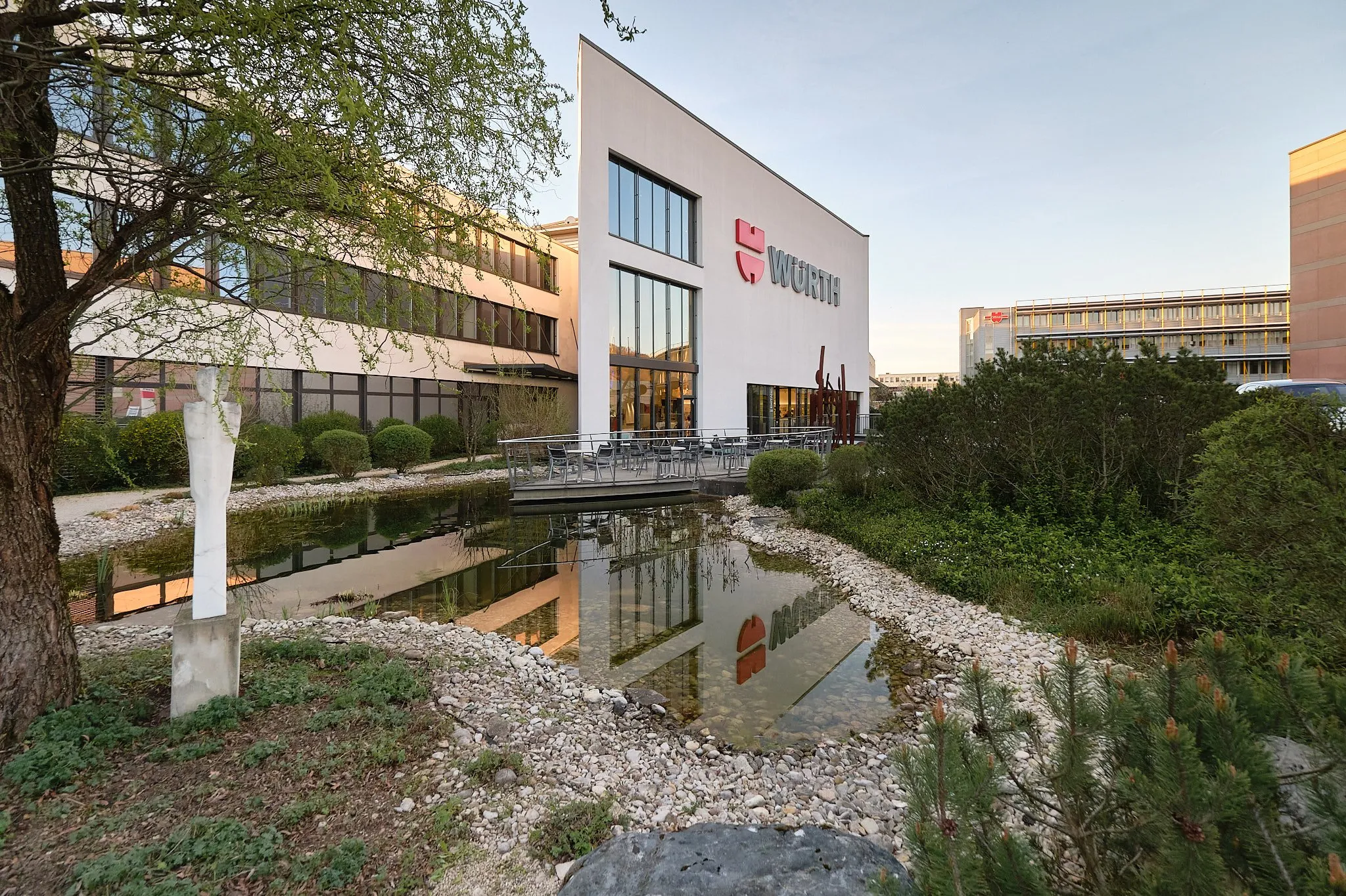 Edificio moderno de color blanco con el logotipo de Würth reflejado en un estanque rectangular, rodeado de vegetación y una escultura blanca.