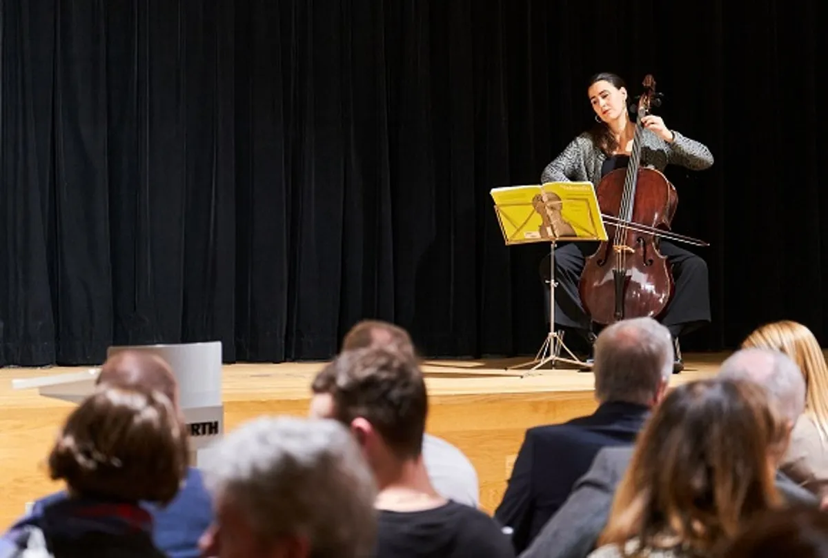 Un violonchelista actúa en un escenario de madera ante una audiencia sentada, con un telón negro de fondo.