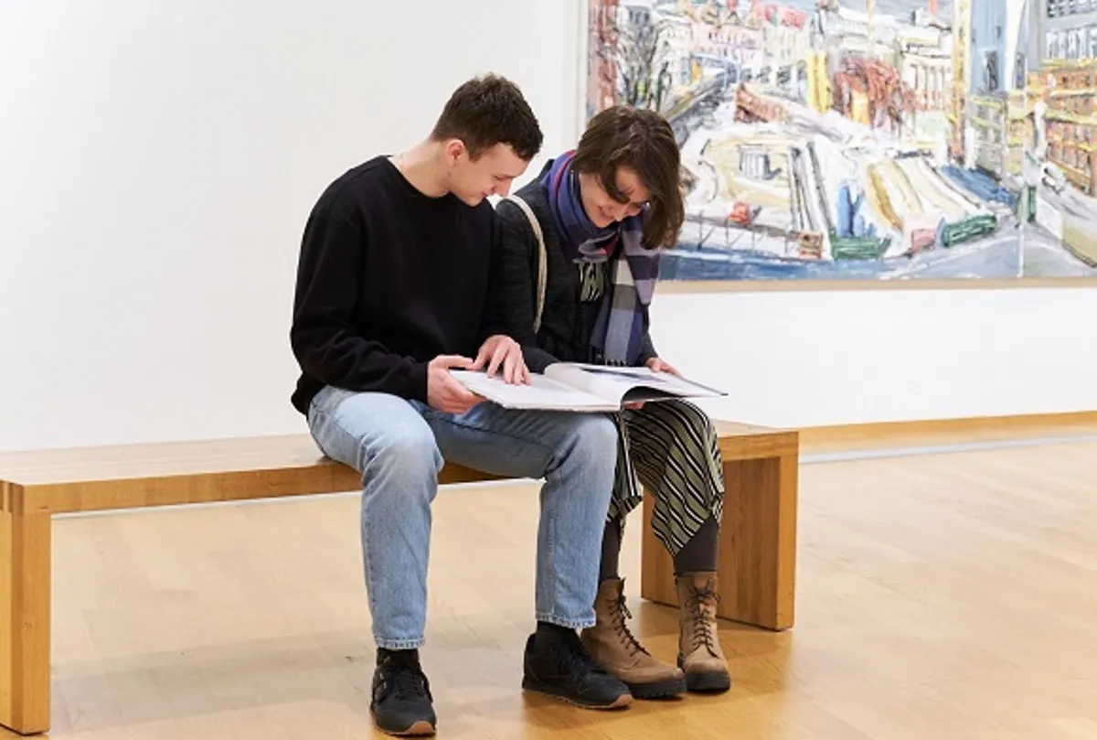 Dos personas sentadas en un largo banco de madera en una galería luminosa, mirando un libro abierto, con un gran cuadro colorido colgado en la pared blanca detrás de ellos.