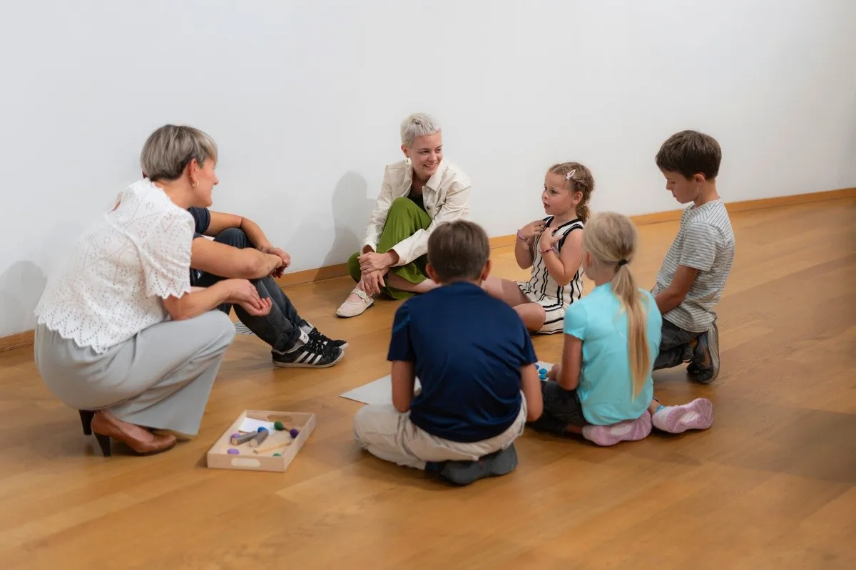 Adultos y niños sentados en círculo sobre un suelo de madera en una galería de paredes blancas durante un taller educativo.