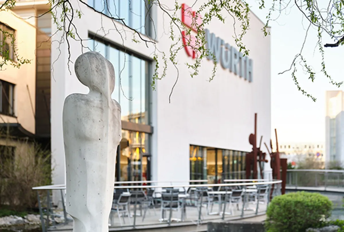 Vista exterior del edificio Forum Würth Arlesheim con una gran escultura abstracta de figura humana blanca en primer plano y el logotipo de Würth en la fachada blanca