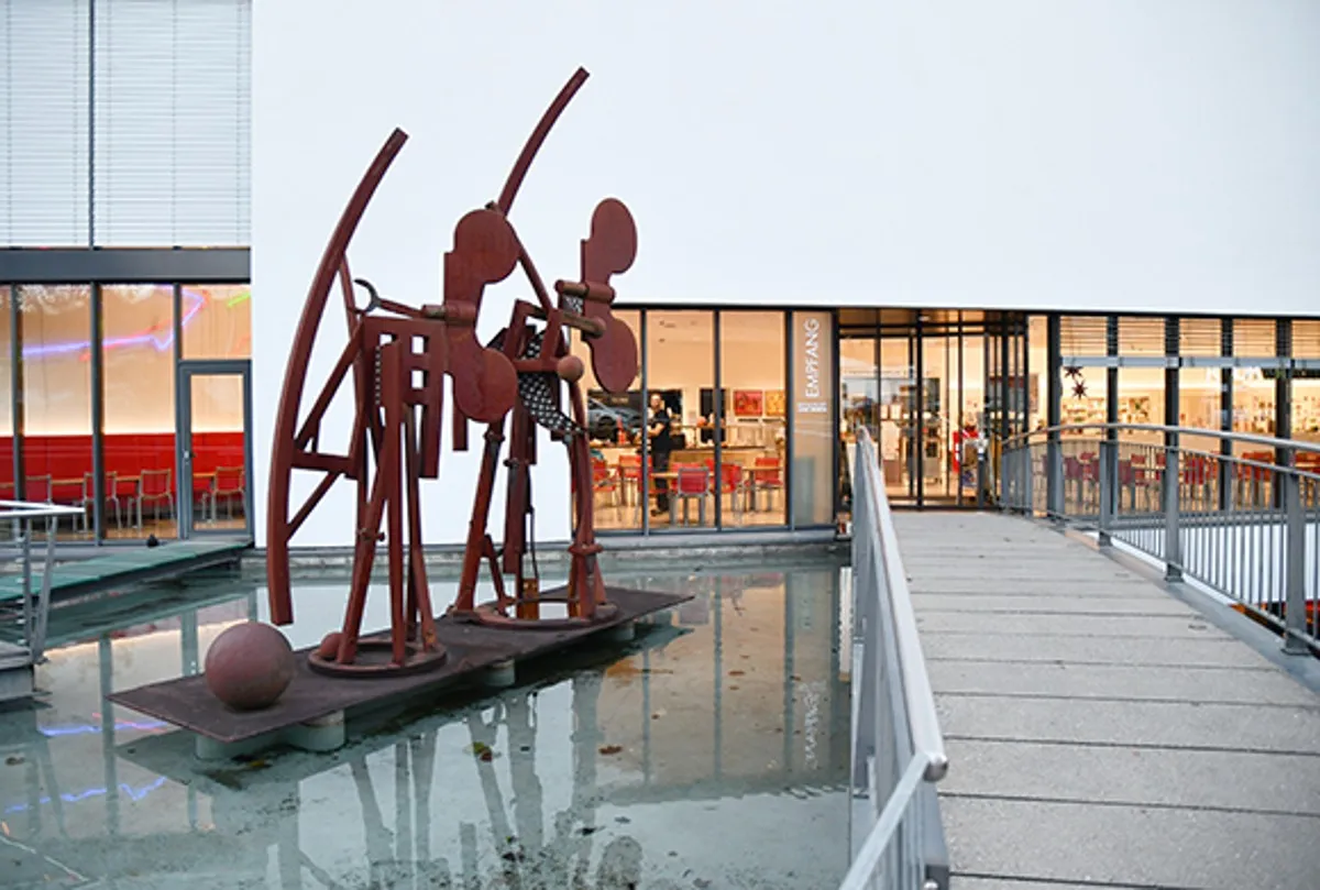 Gran escultura abstracta de metal de color rojizo-marrón situada en una fuente poco profunda al aire libre, frente a la entrada de un edificio moderno con paredes de cristal.