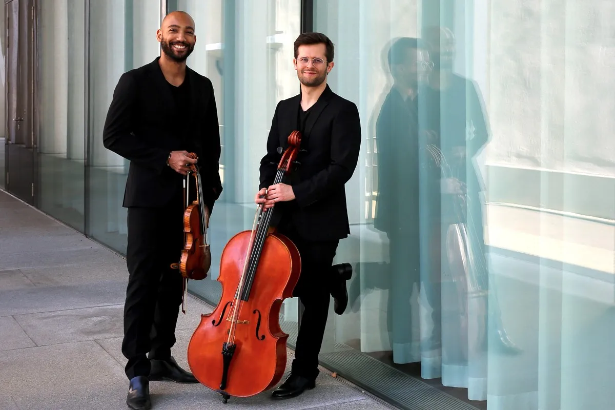Dos hombres con trajes negros están de pie, uno al lado del otro, sosteniendo un violín y un violonchelo frente a una pared de cristal, con sus reflejos visibles en el vidrio.