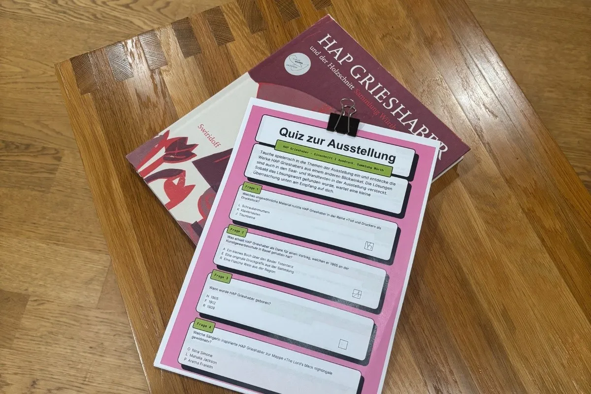 Una hoja de cuestionario titulada "Quiz zur Ausstellung" sujeta a un portapapeles rosa, apoyada sobre una mesa de madera con un catálogo de la exposición de Hap Grieshaber debajo.