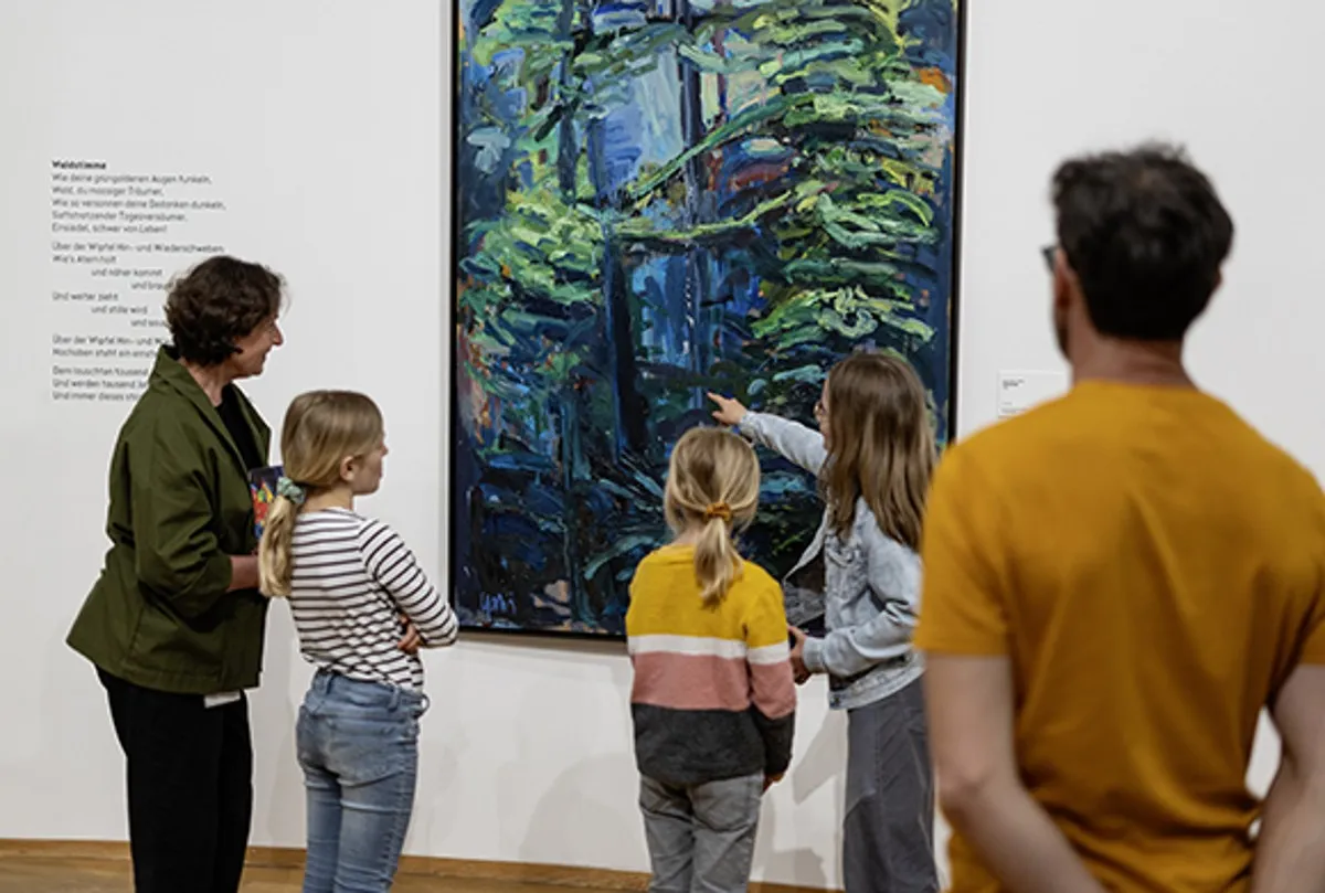 Adultos y niños de pie en una sala de galería blanca observan una gran pintura de un bosque azul verdoso en la pared