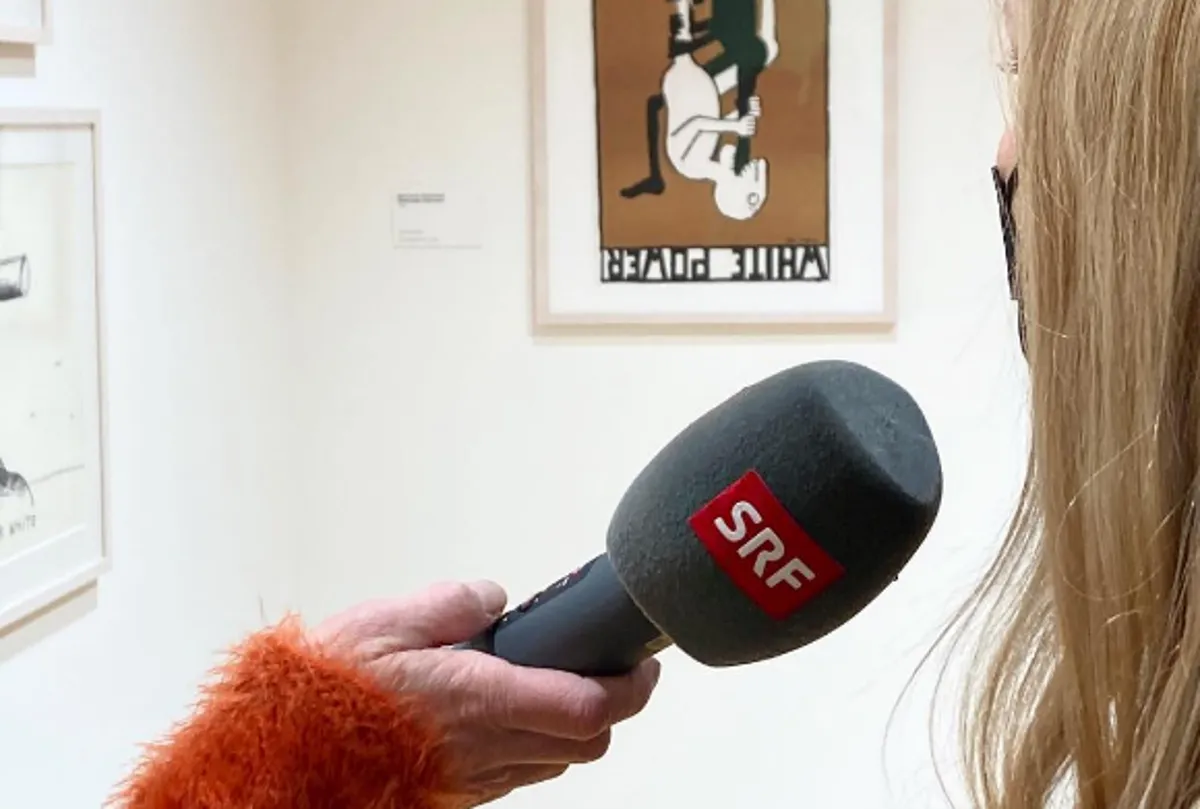 Una mano sosteniendo un micrófono de SRF durante una entrevista frente a obras de arte enmarcadas en la pared blanca de una galería