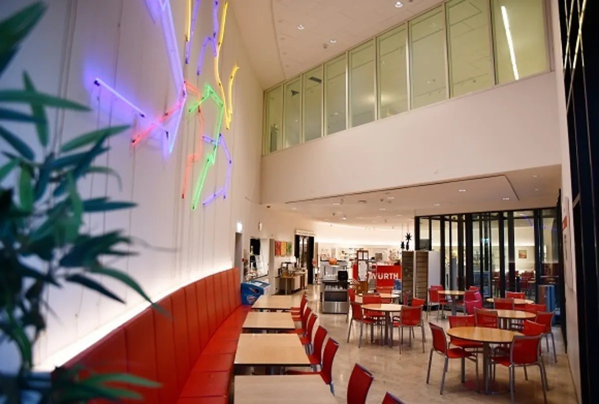 Interior del Forum Würth Arlesheim que muestra una zona de asientos con sillas y mesas rojas, un largo banco rojo a lo largo de una pared con una colorida instalación de luces de neón y un altillo con ventanales de cristal.