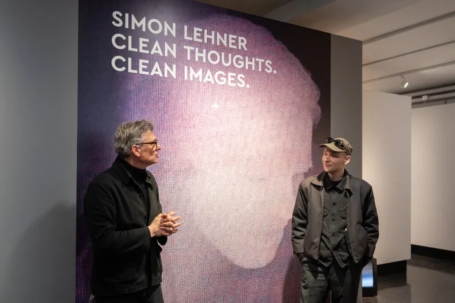 Dos hombres de pie en un espacio de exposiciones frente a un gran cartel morado con texto blanco