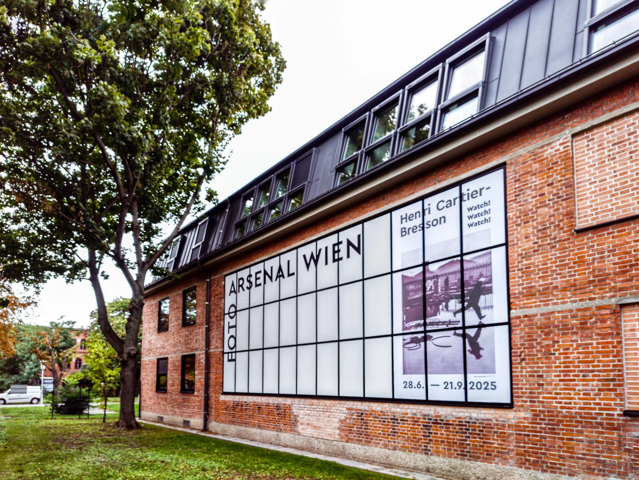 Rood bakstenen gebouw met de tekst 'FOTO ARSENAL WIEN' op een groot glazen paneel, omgeven door groen gras en bomen.