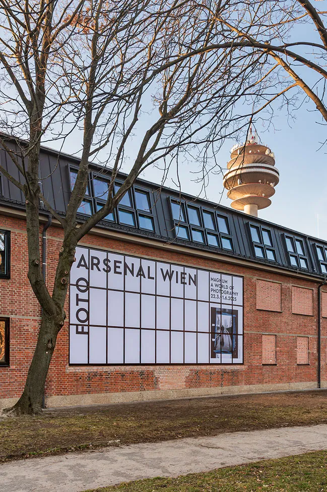 Bakstenen gebouw met groot wit bord waarop 'FOTO ARSENAL WIEN' staat en tentoonstellingsdetails, met de Wiener Stadtturm zichtbaar op de achtergrond.
