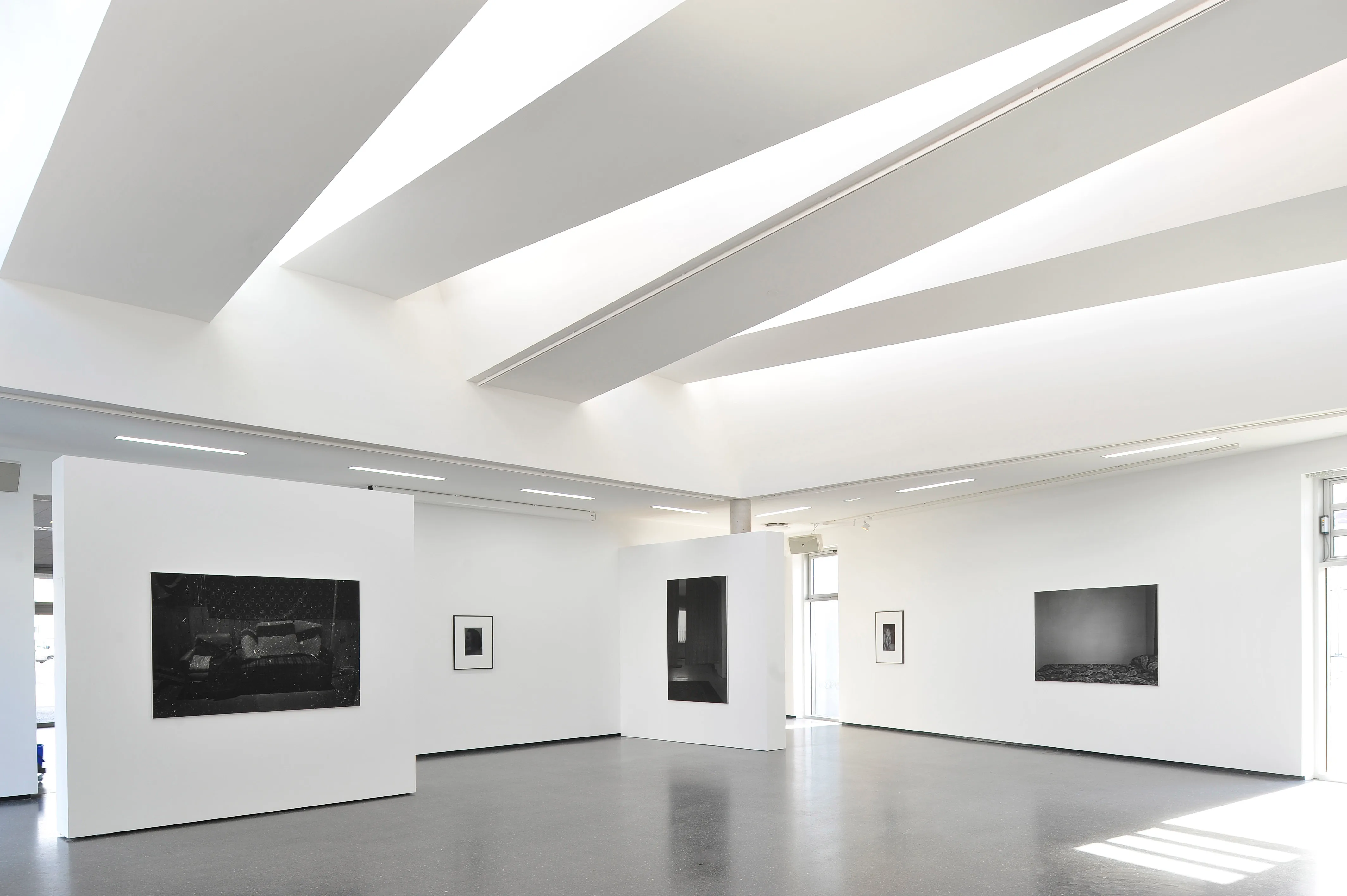 Interior de galería blanco brillante con tragaluces inclinados y fotografías en blanco y negro expuestas en paredes blancas independientes