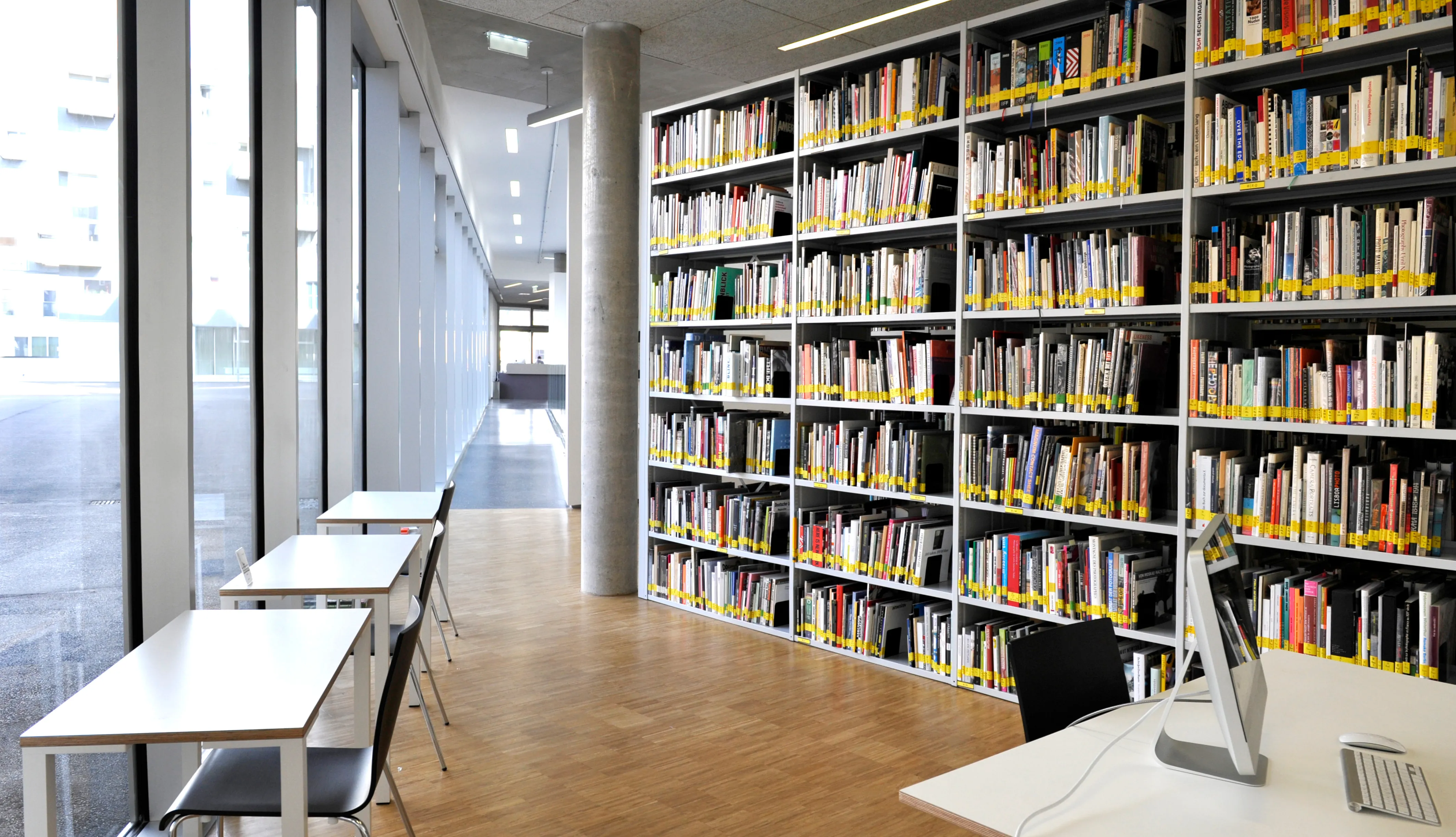 Vista interior de la biblioteca Fotohof con estanterías blancas altas llenas de libros, mesas de lectura con sillas y grandes ventanales a lo largo de la pared izquierda
