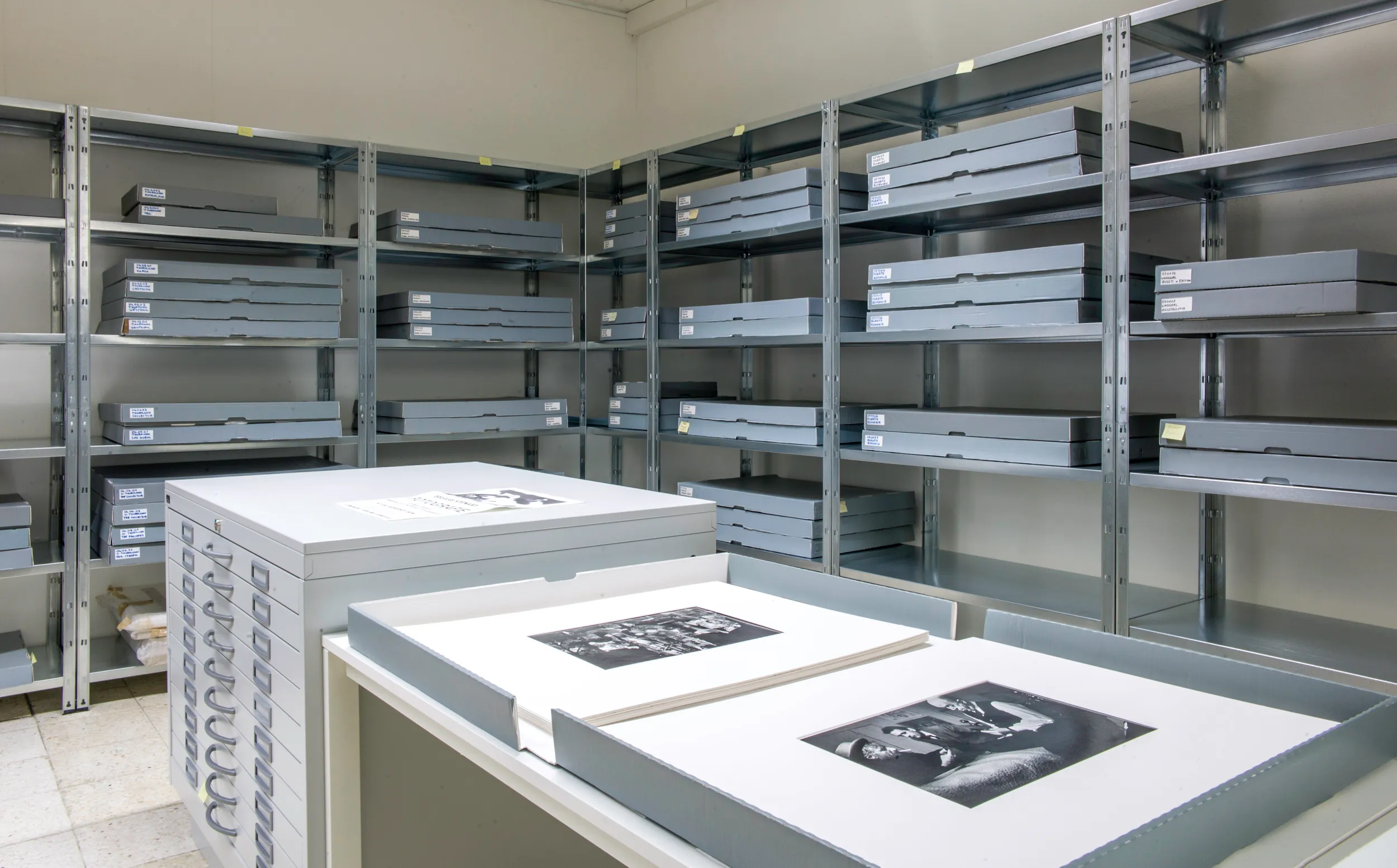 Sala de archivos con estanterías metálicas llenas de cajas de almacenamiento grises y una mesa que muestra cajas de archivo abiertas con fotografías en blanco y negro