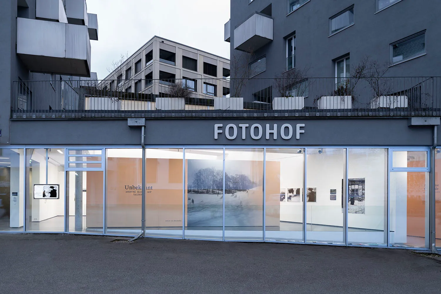 Vista exterior a nivel de calle del centro de fotografía FOTOHOF, que muestra un amplio escaparate de cristal con el nombre del lugar en grandes letras blancas encima, con exposiciones de la galería visibles a través de las ventanas y modernos edificios de apartamentos a ambos lados.