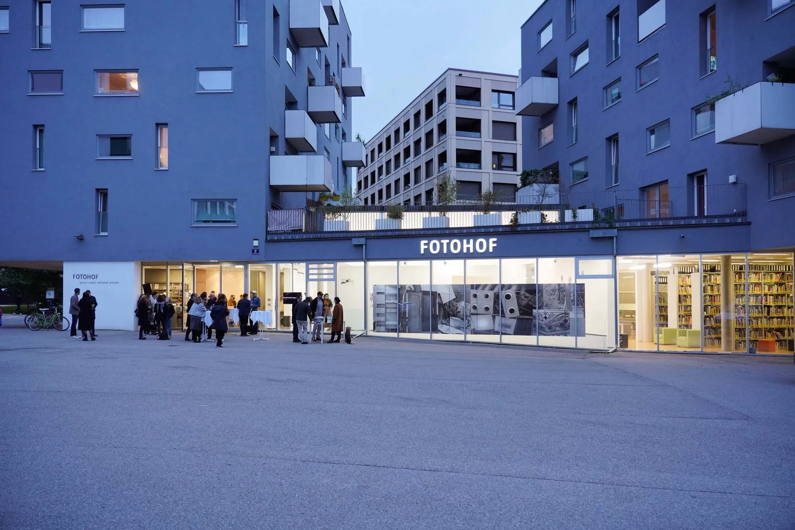 Vista exterior del edificio FOTOHOF al atardecer, con gente reunida frente a la entrada y grandes ventanales que muestran el interior.