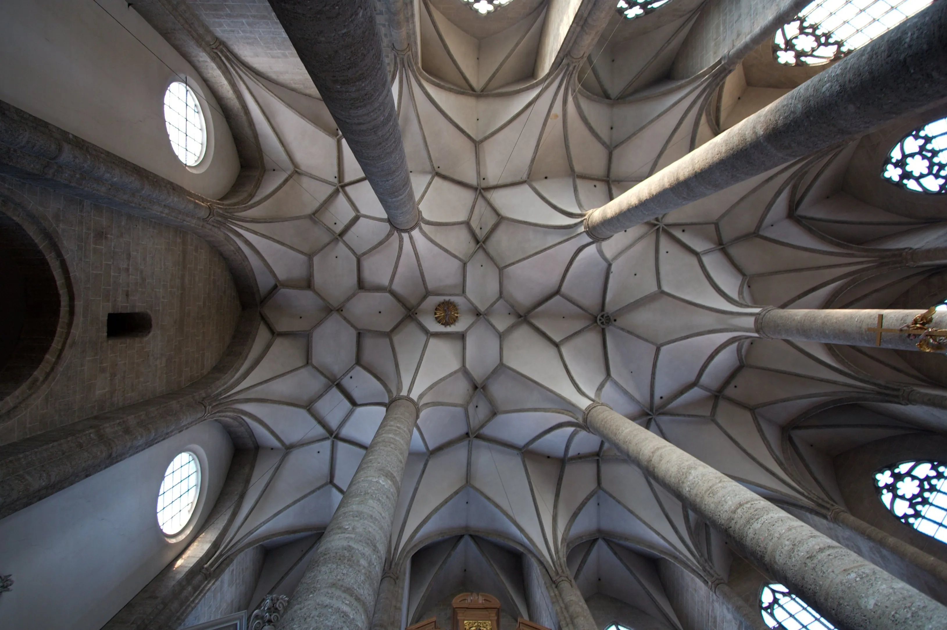 Upward view of the ribbed vaulted ceiling inside the Franciscan Church, with stone columns rising to meet an intricate patterned stone ceiling and circular windows along the walls.