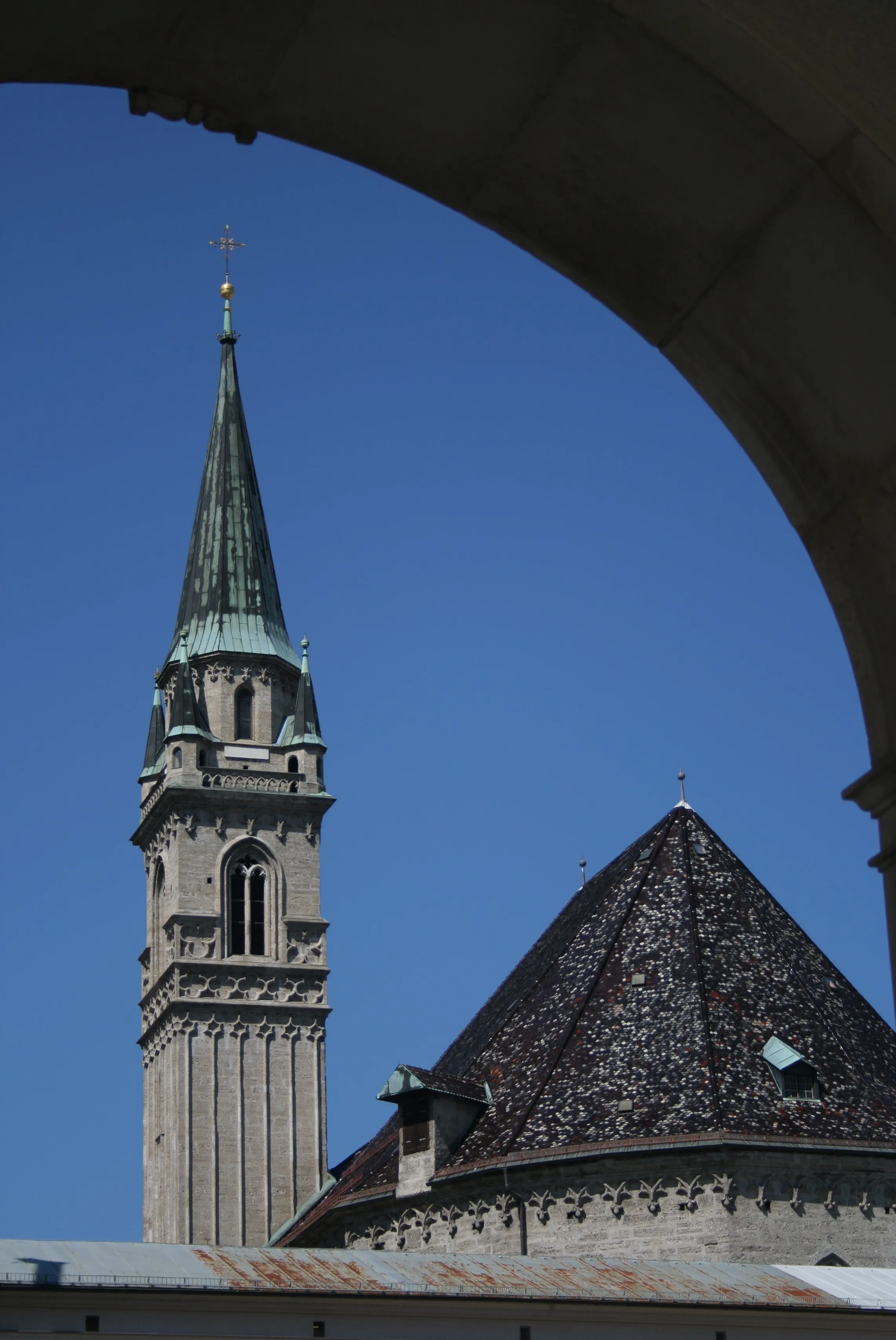 Tall stone church tower with a green spire and cross, next to a dark tiled roof, framed by a stone archway against a clear blue sky.
