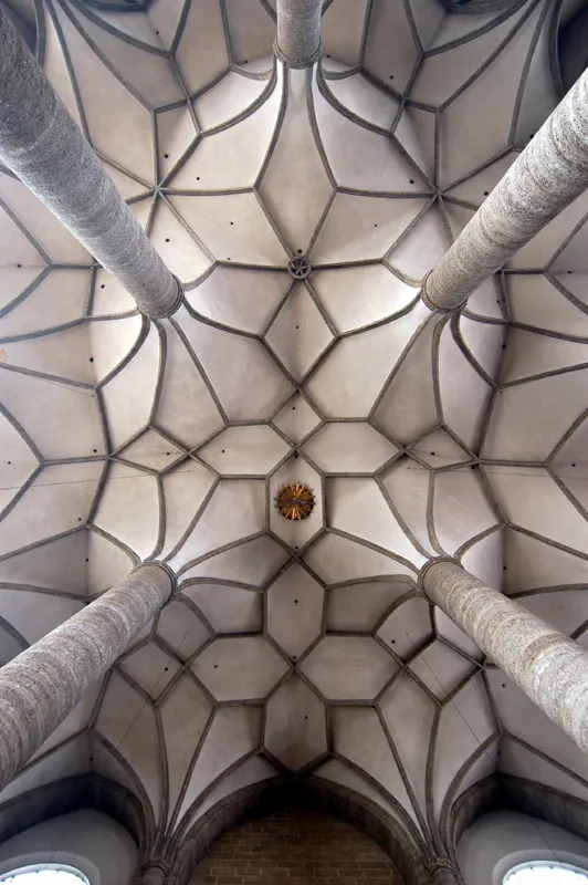 Looking up at an intricate Gothic ribbed vault ceiling with four large stone columns and a geometric stone rib network inside Franciscan Church Salzburg