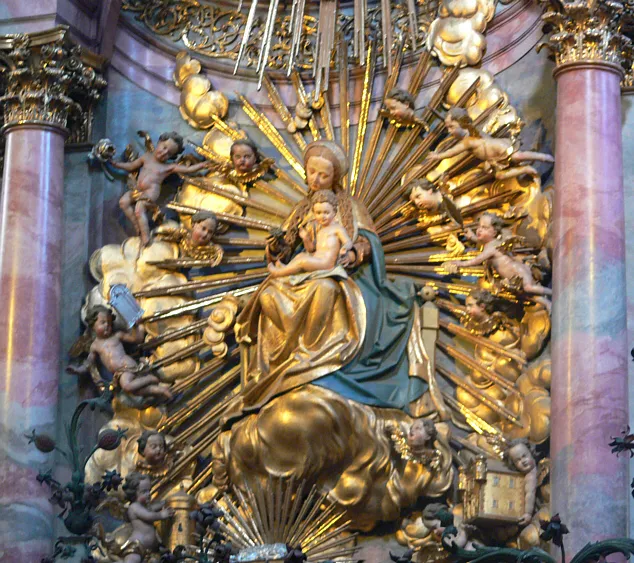 Ornate gilded baroque high altar sculpture depicting the Virgin Mary holding the Christ Child, surrounded by cherubs and radiating golden sunburst rays, flanked by pink marble columns.
