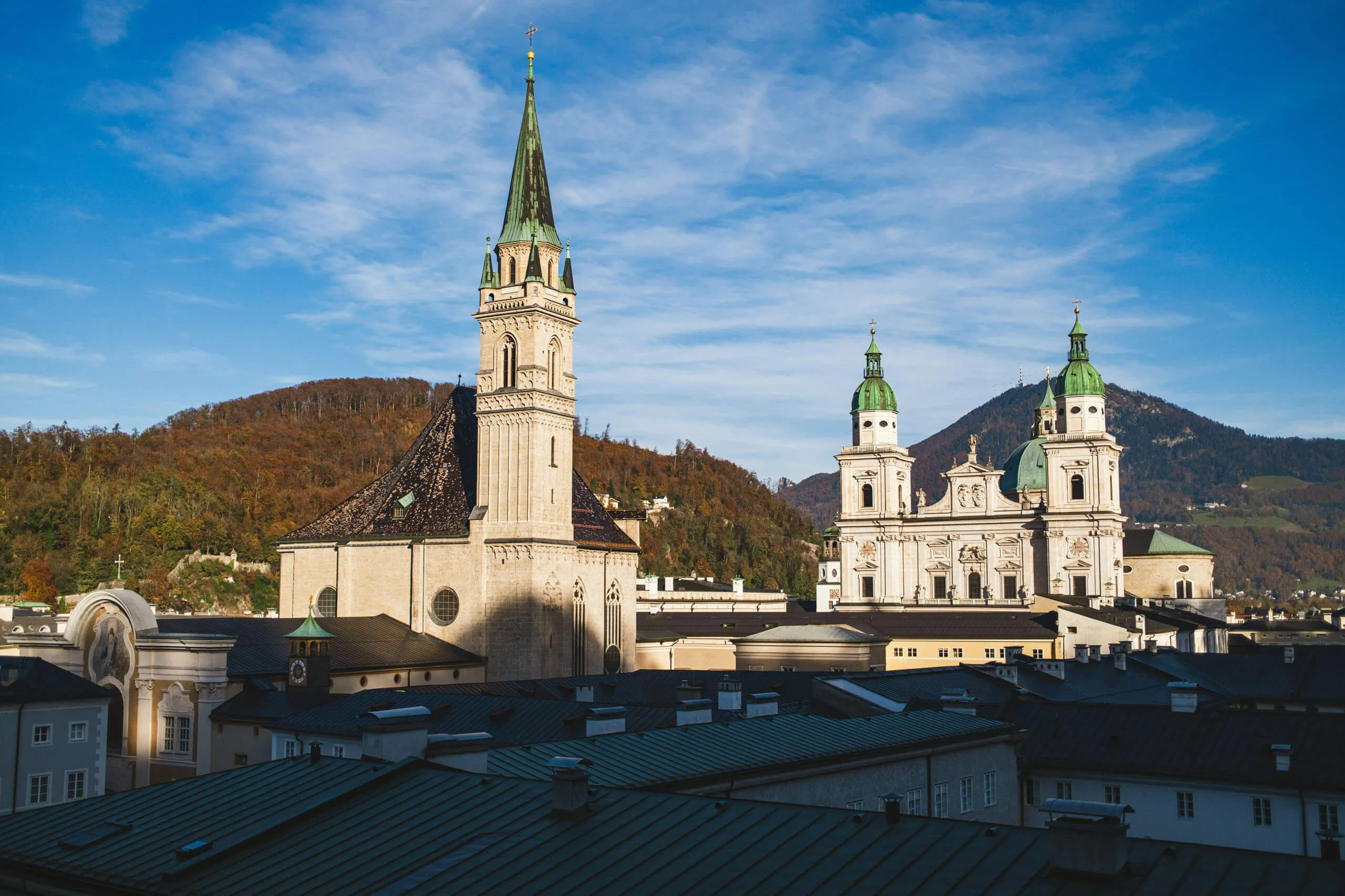 A cityscape view featuring the tall spire of the Franciscan Church and the twin towers of Salzburg Cathedral against a backdrop of mountains and blue sky.