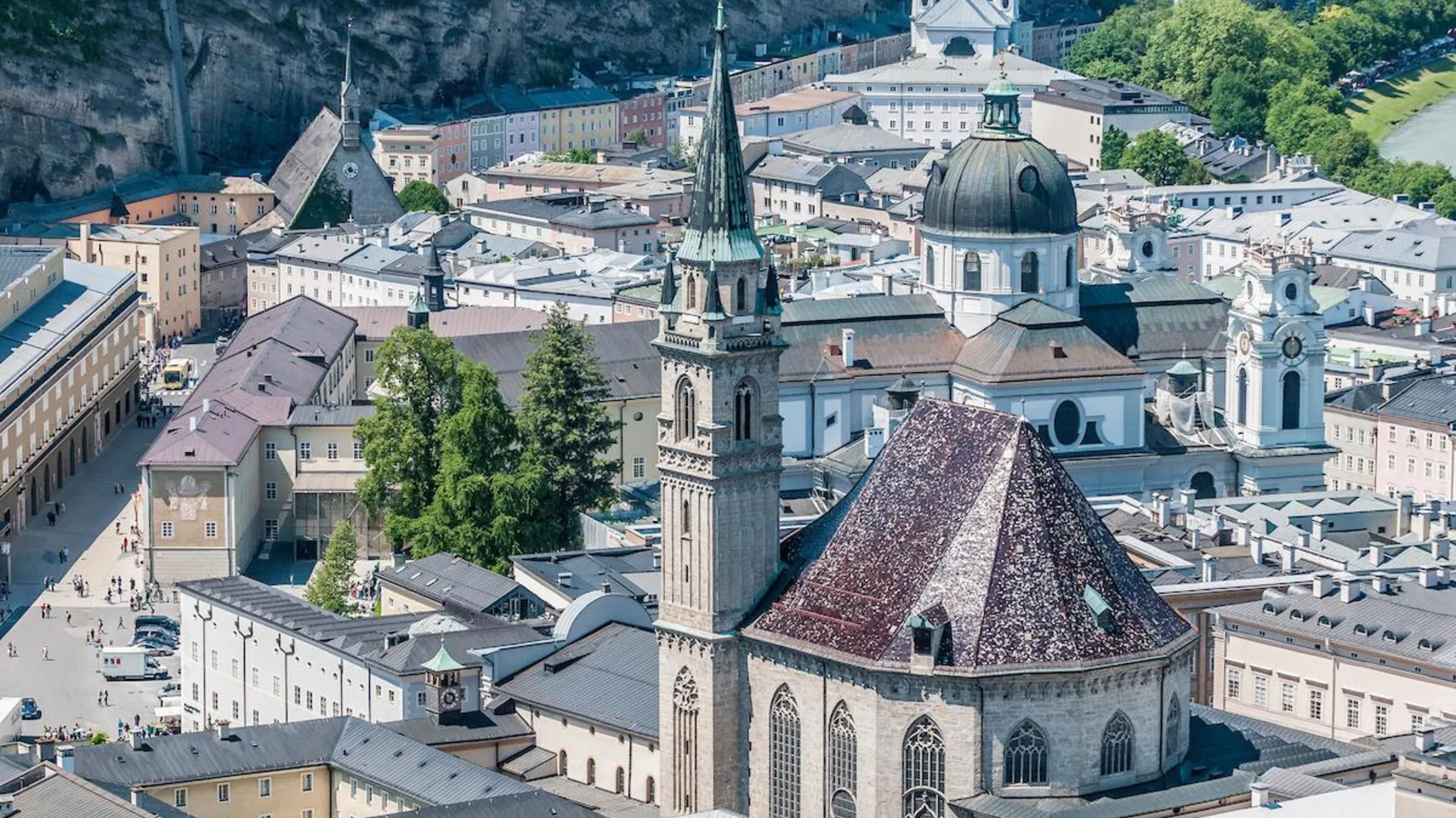 Aerial view of the Franciscan Church in Salzburg showing its tall green spire and reddish-brown tiled polygonal apse, surrounded by the historic city center with the Salzburg Cathedral dome nearby.