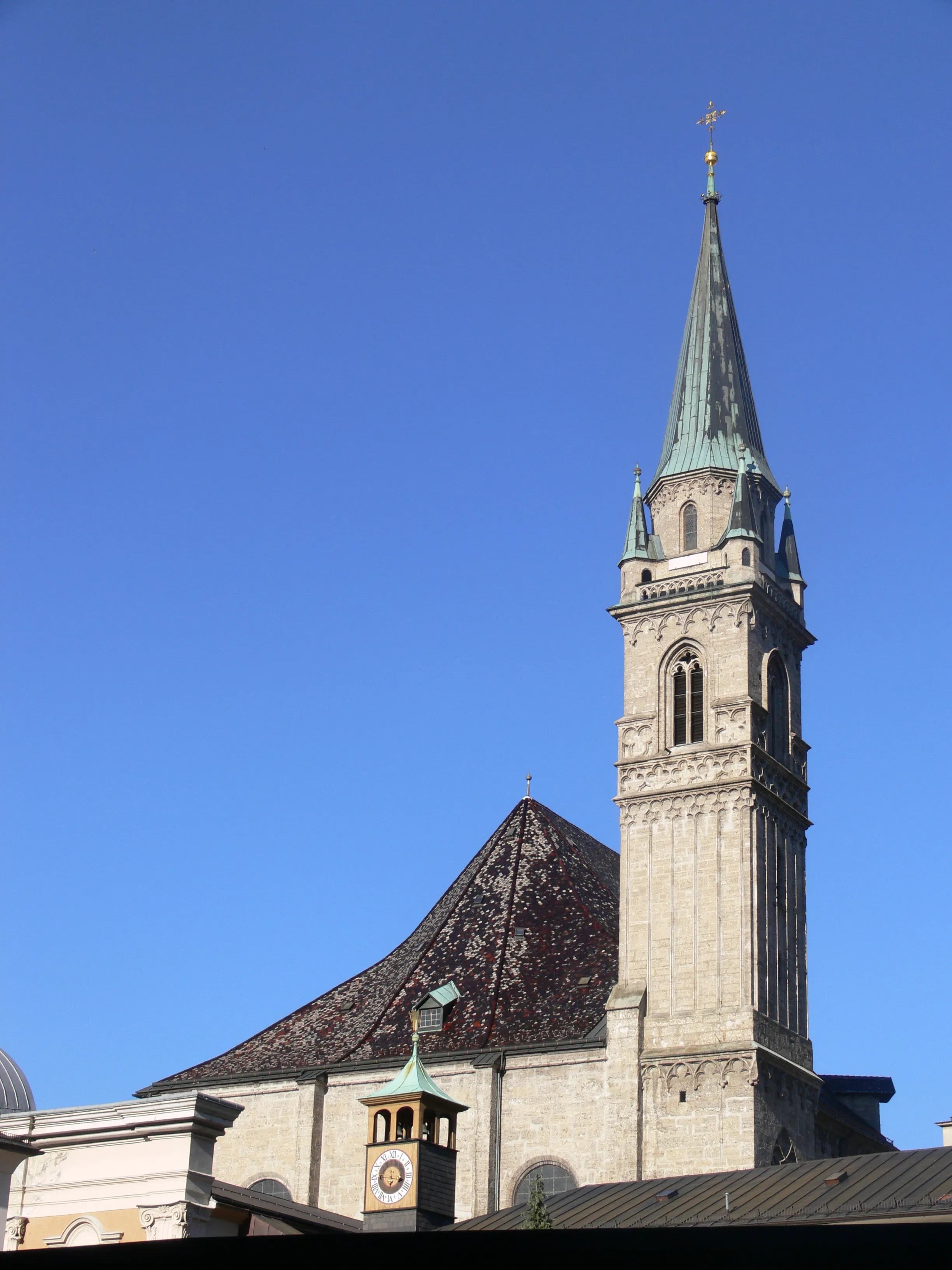 Tall stone church tower with green copper spire and cross against blue sky, with dark tiled roof and small clock tower below