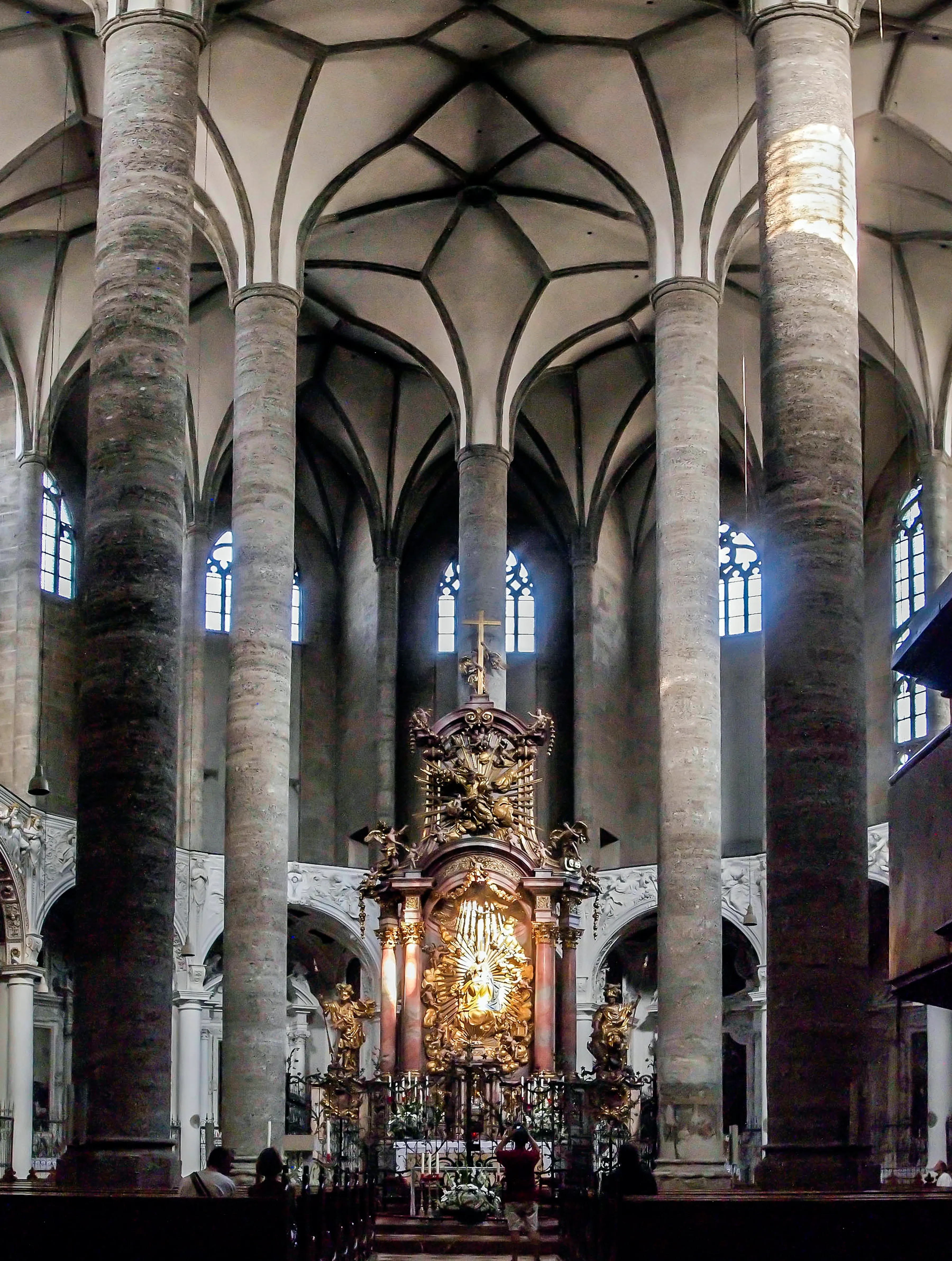 Interior of the Franciscan Church in Salzburg showing tall stone columns, a ribbed Gothic vaulted ceiling, and an ornate gilded high altar with a cross at the far end