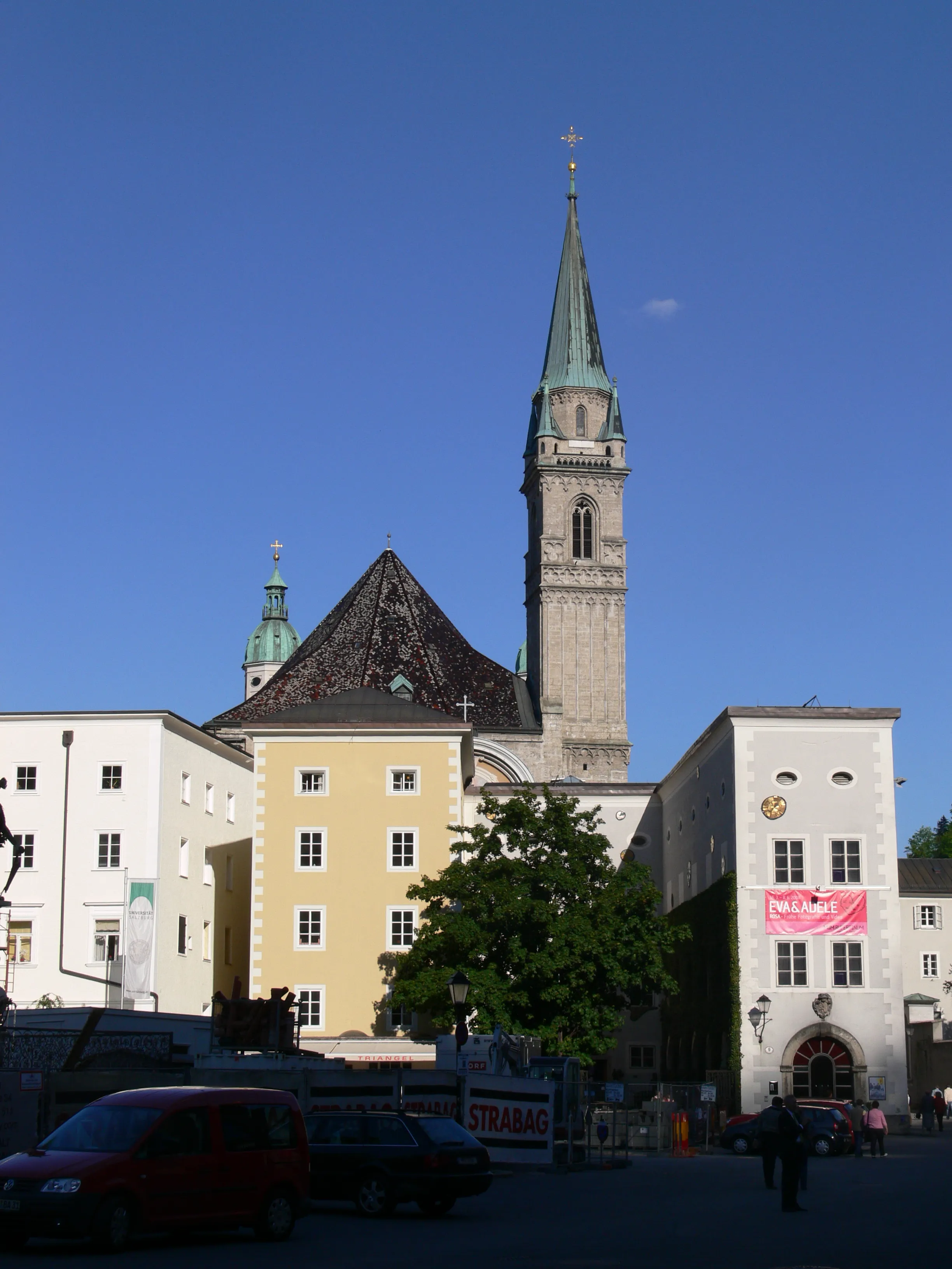 Hoge kerktoren met een groene koperen spits en kruis die boven witte en gele gebouwen in Salzburg uitsteekt, met geparkeerde auto's en voetgangers op het plein op de voorgrond.