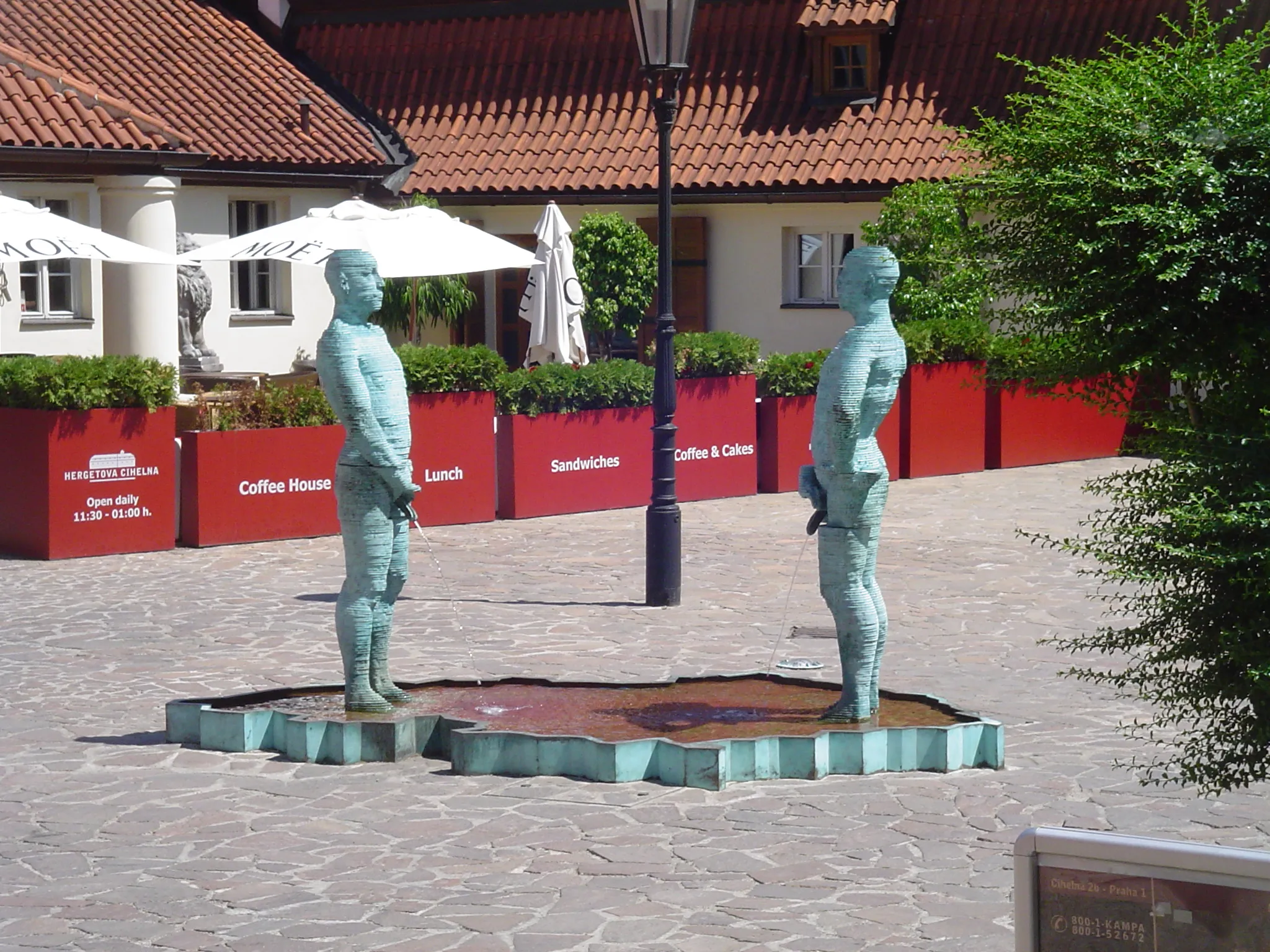 Two bronze statues of Franz Kafka in a courtyard with red planters and a building with a tiled roof