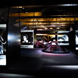 Interieur van een museum met donkere verlichting, houten balken en verlichte vitrines