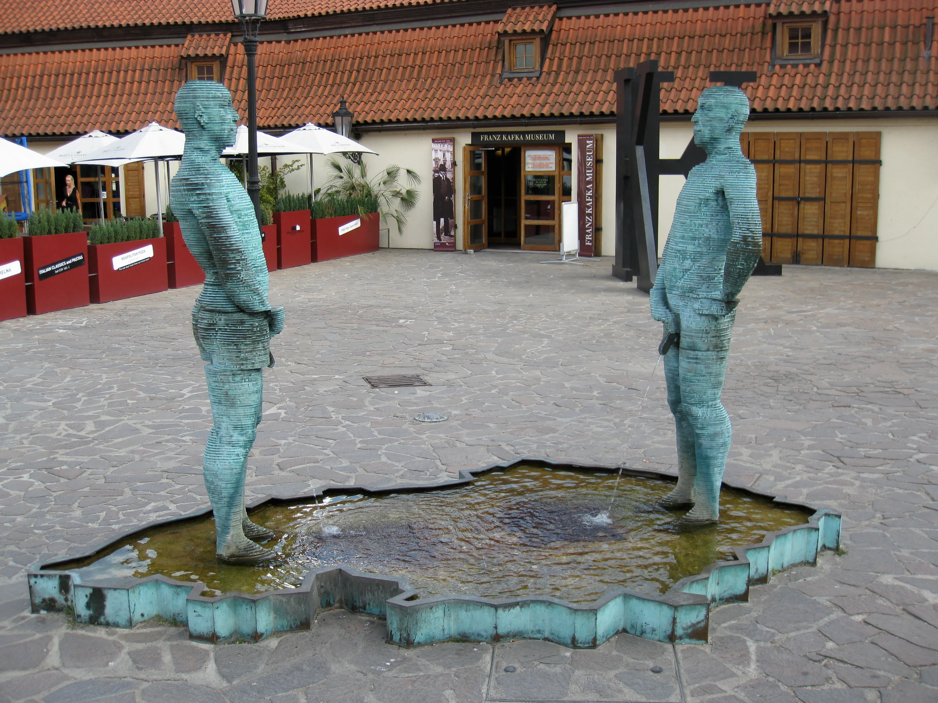 Twee groene bronzen beelden van staande figuren die tegenover elkaar staan boven een waterpartij buiten een gebouw met een rood pannendak
