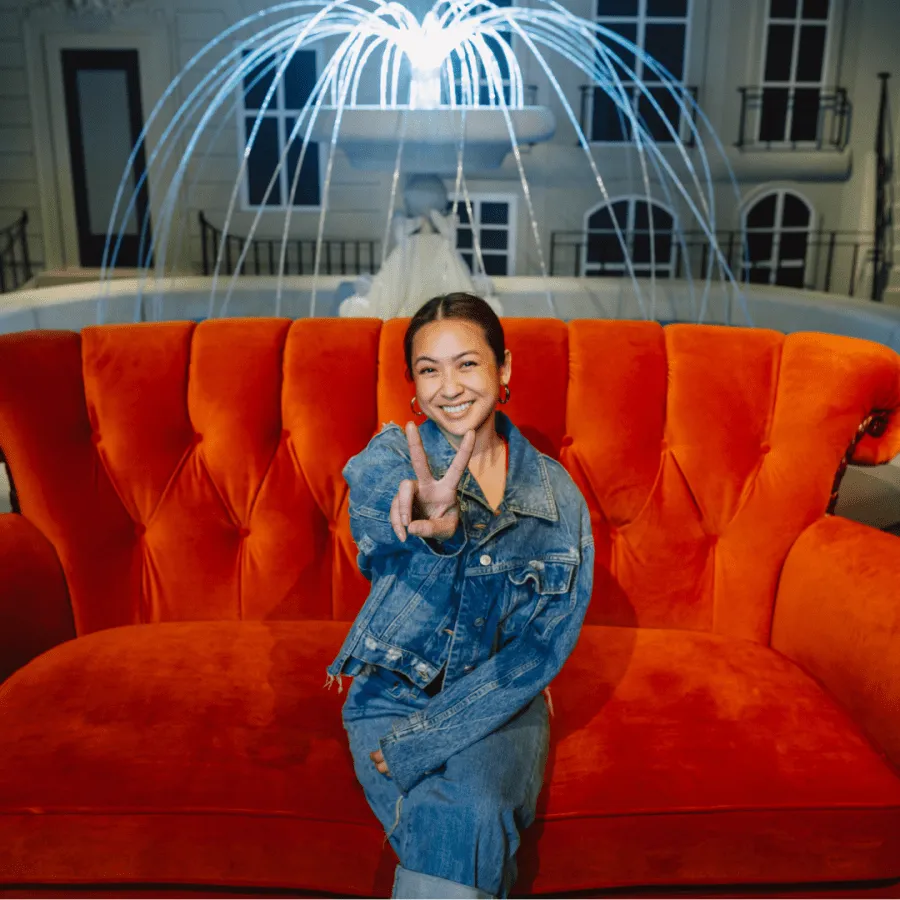 A woman in a denim jacket sitting on a bright orange tufted sofa, making a peace sign, with a fountain and building facade behind her.