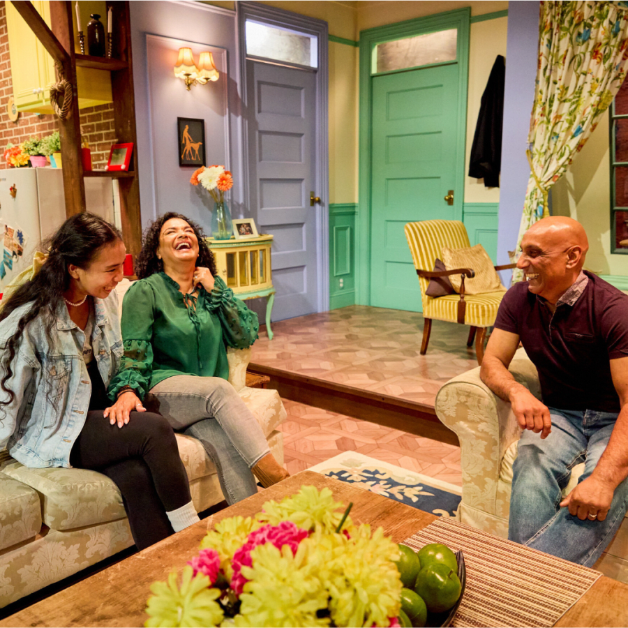 Three people laughing in a recreated living room with a purple door and floral couch