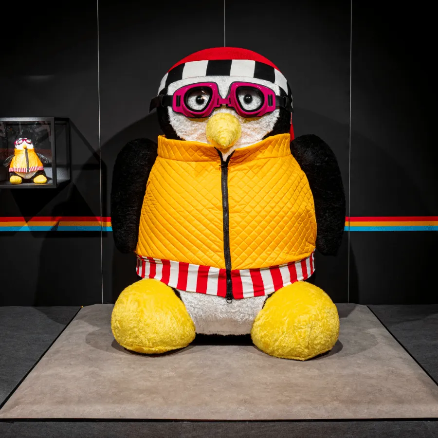 Large penguin figure wearing a yellow vest, red and white striped pants, and pink goggles