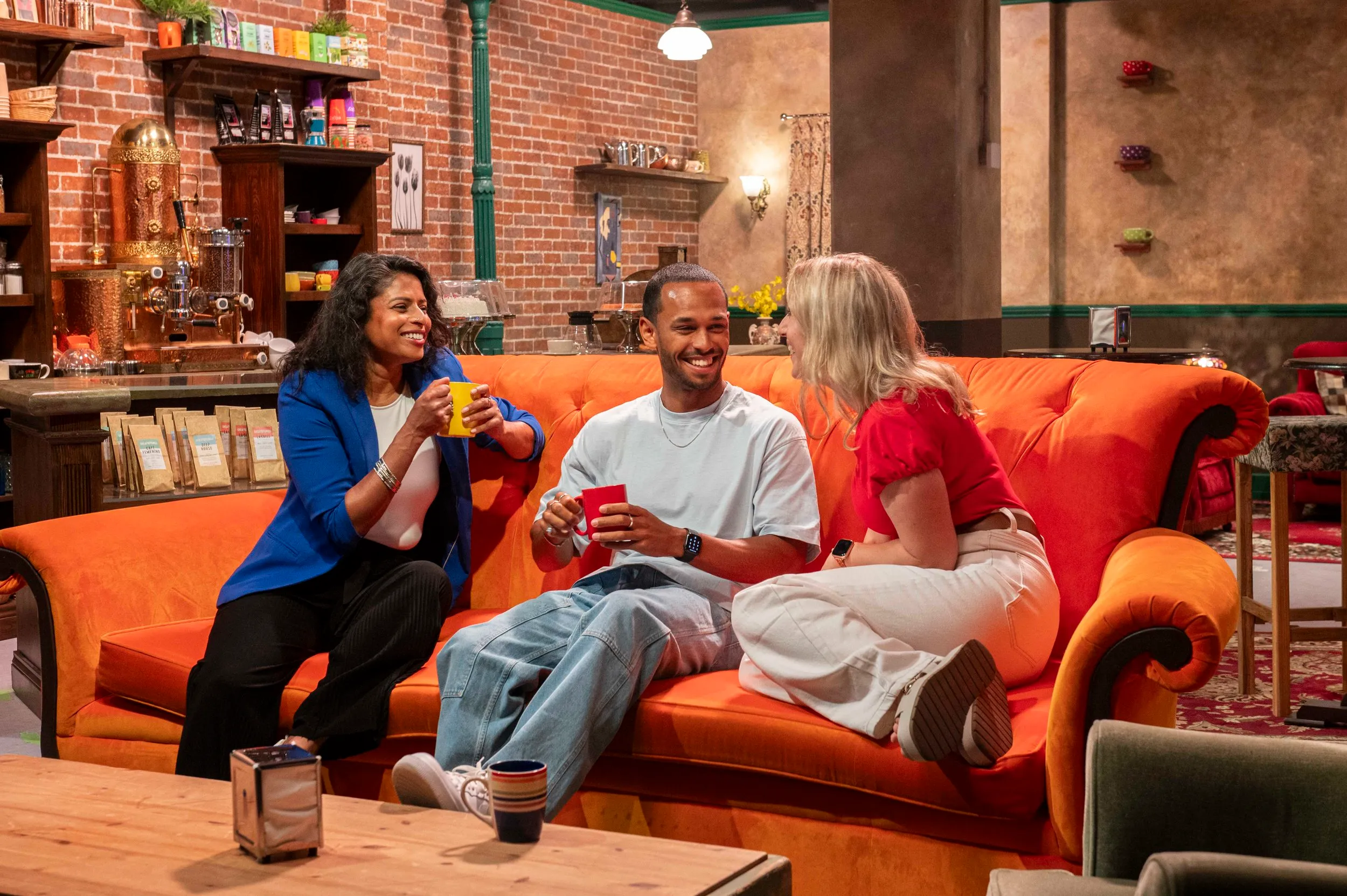 Drie mensen die op de oranje bank zitten in de Central Perk-set van FRIENDS™