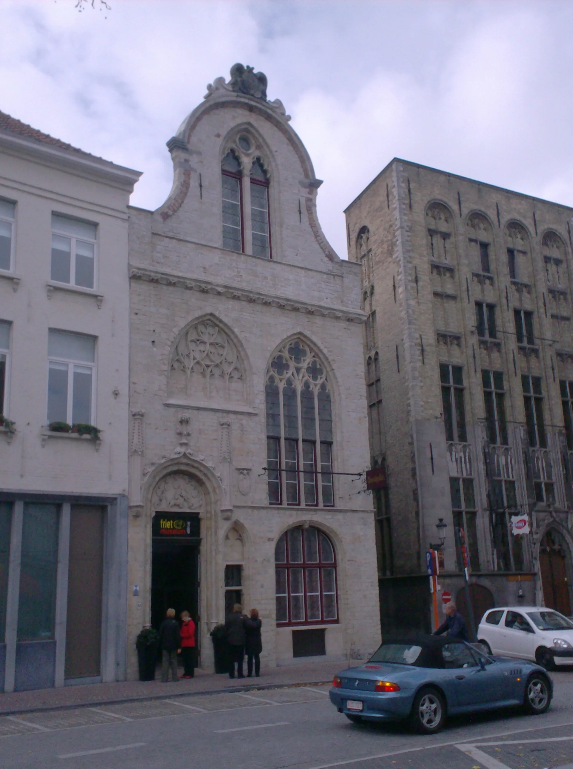 Fachada de piedra medieval del edificio Saaihalle en Brujas con ventanas de arco gótico, hastial escalonado y personas de pie en la entrada