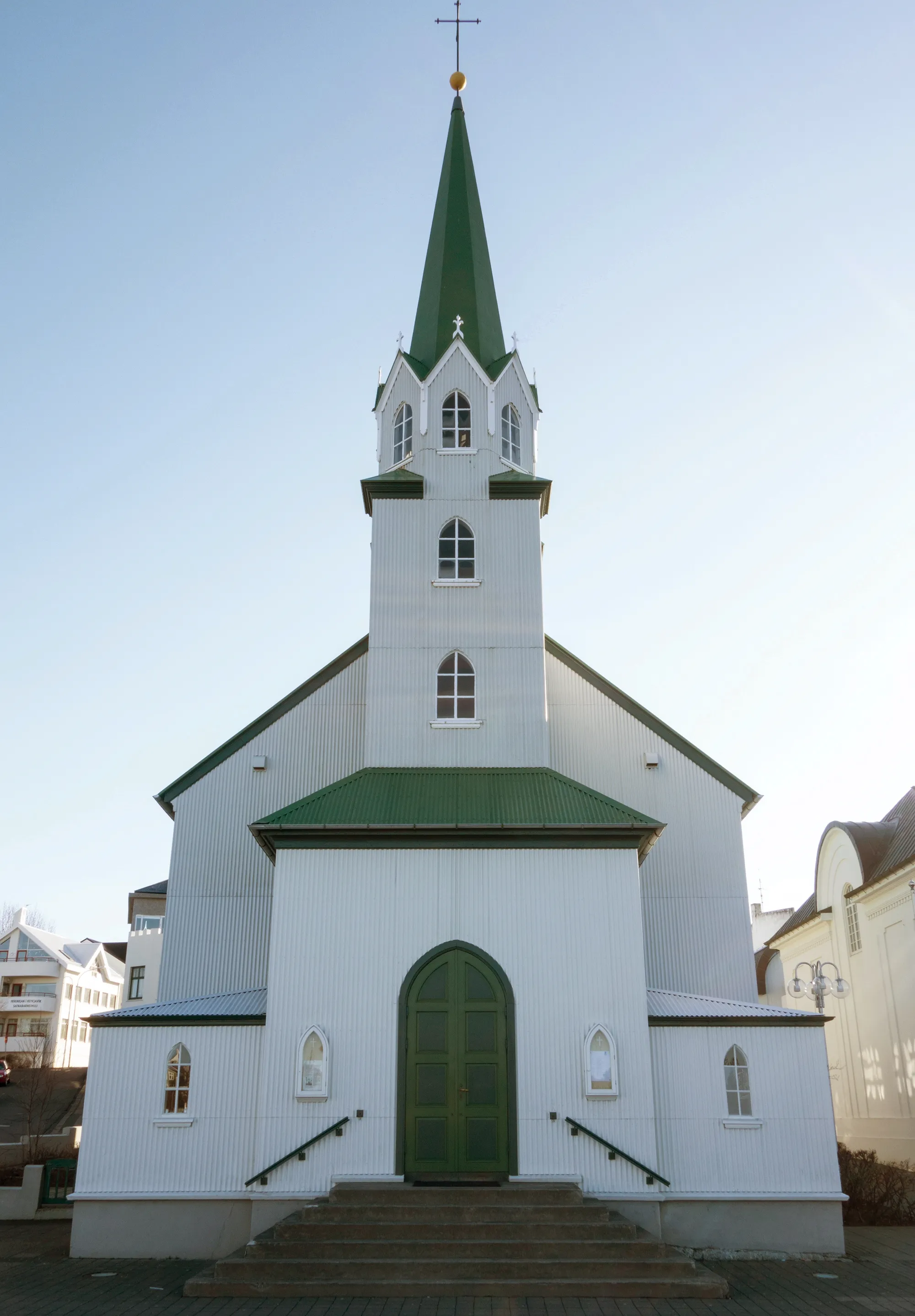 Iglesia blanca con tejado verde y alta aguja verde rematada con una cruz, fachada frontal con puerta arqueada verde y escalones.