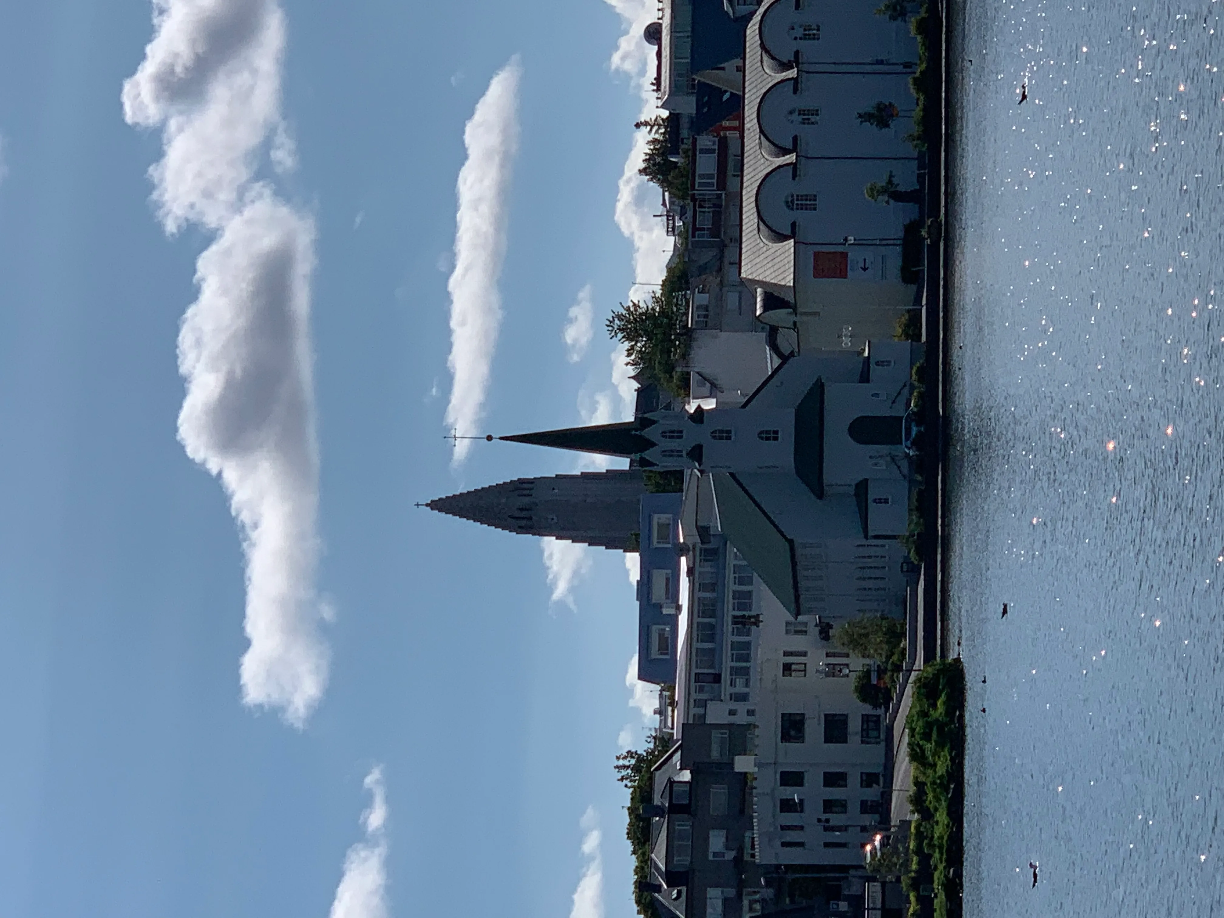 Iglesia blanca con una torre oscura y techo verde a orillas de un lago bajo un cielo azul con nubes.