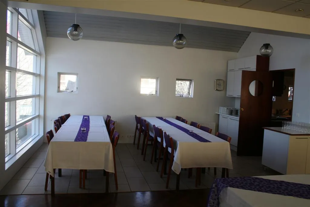 Interior de un salón comunitario con mesas largas cubiertas con manteles blancos y caminos de mesa morados, grandes ventanales a la izquierda y una zona de cocina al fondo.